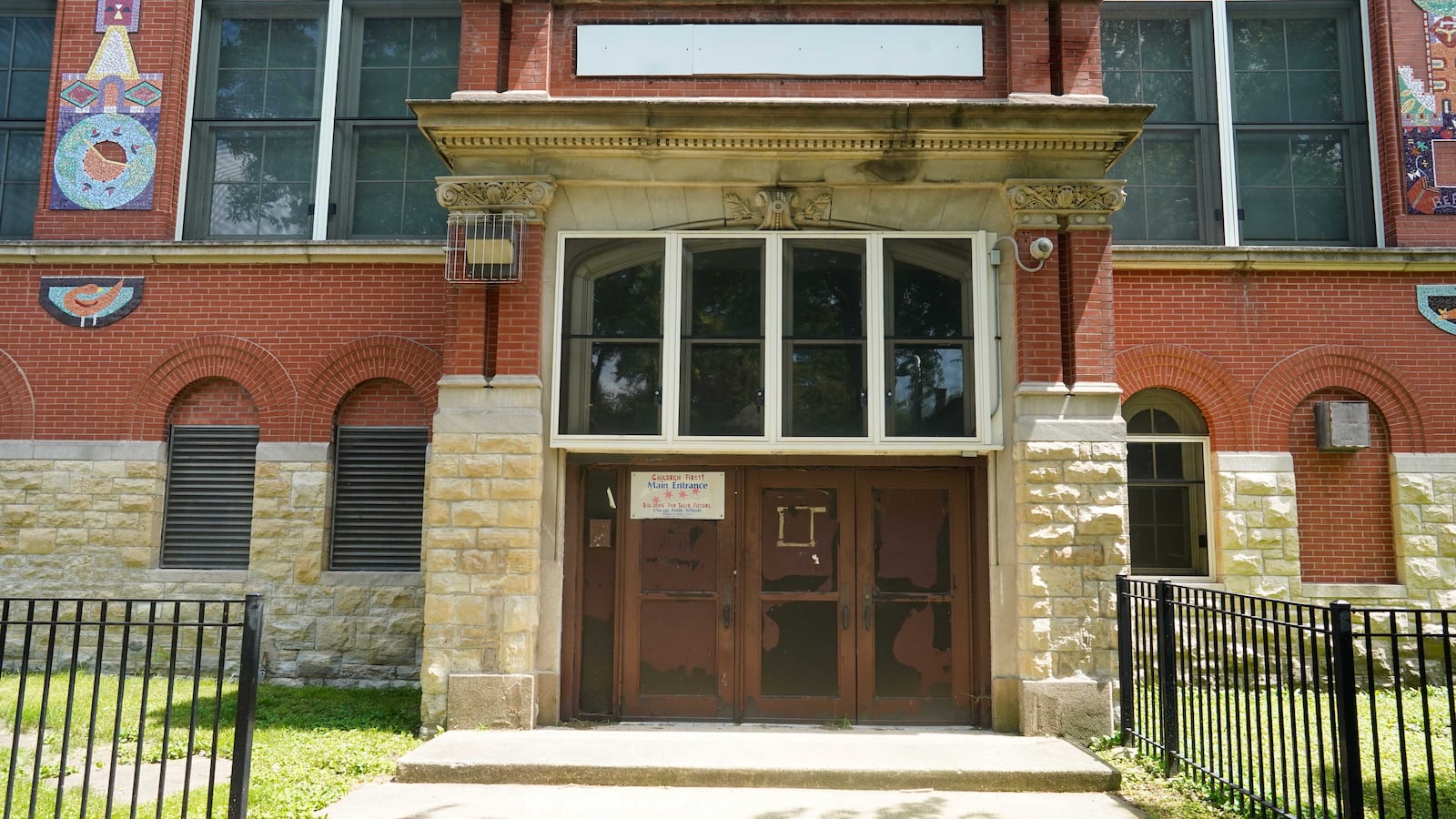 The front entrance of a closed school made of large stone and a wooden door. There are several large windows and a green lawn with a black fence in the foreground.