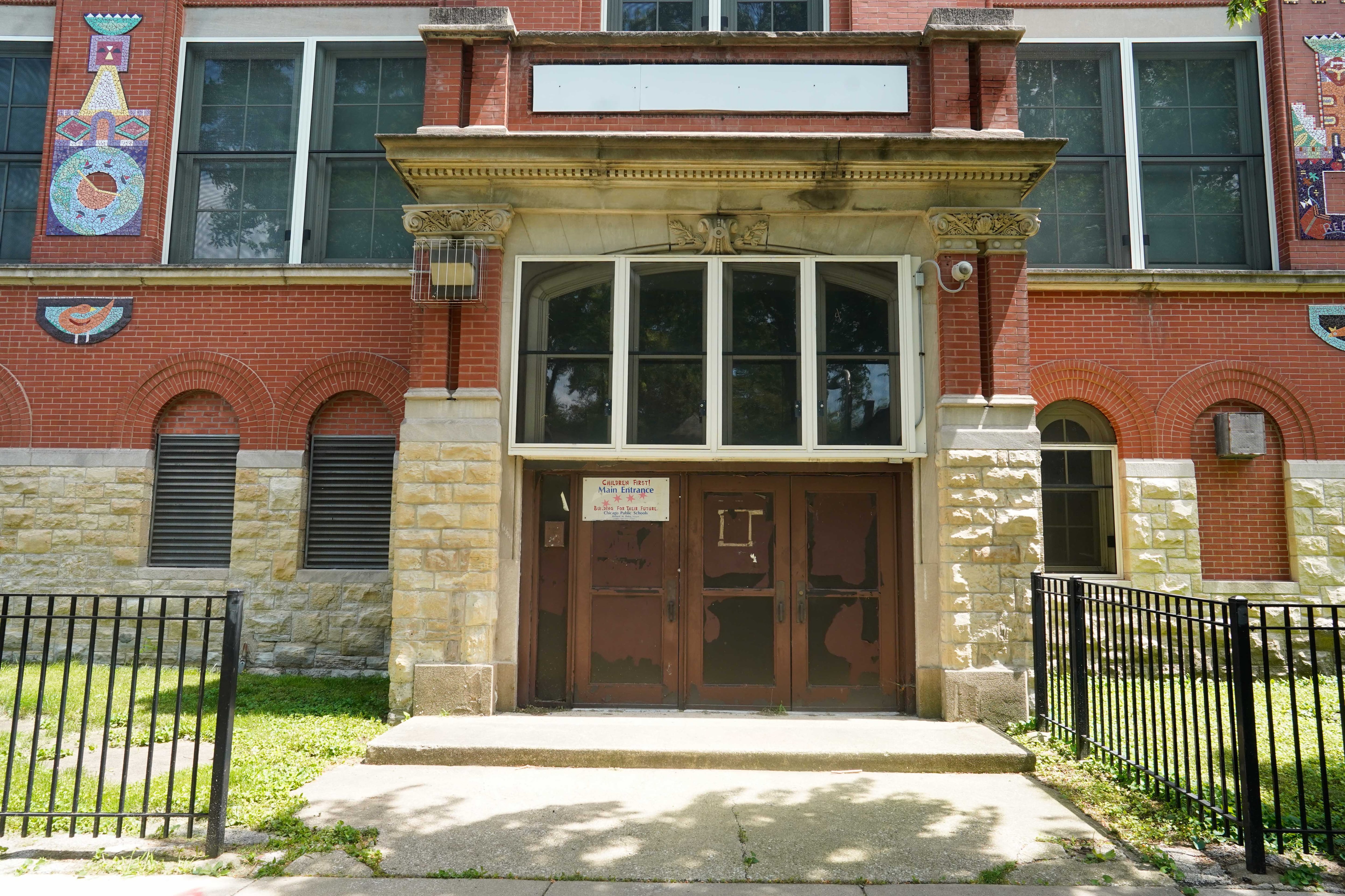 The front entrance of a closed school made of large stone and a wooden door. There are several large windows and a green lawn with a black fence in the foreground.