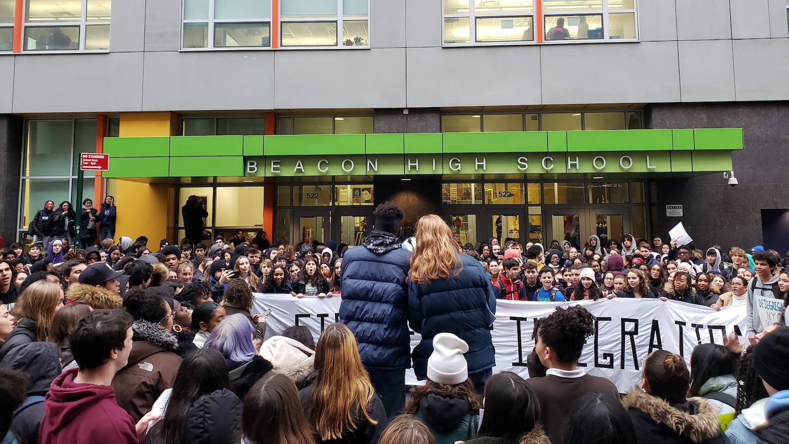 Hundreds of teens gather in front of a high school building.