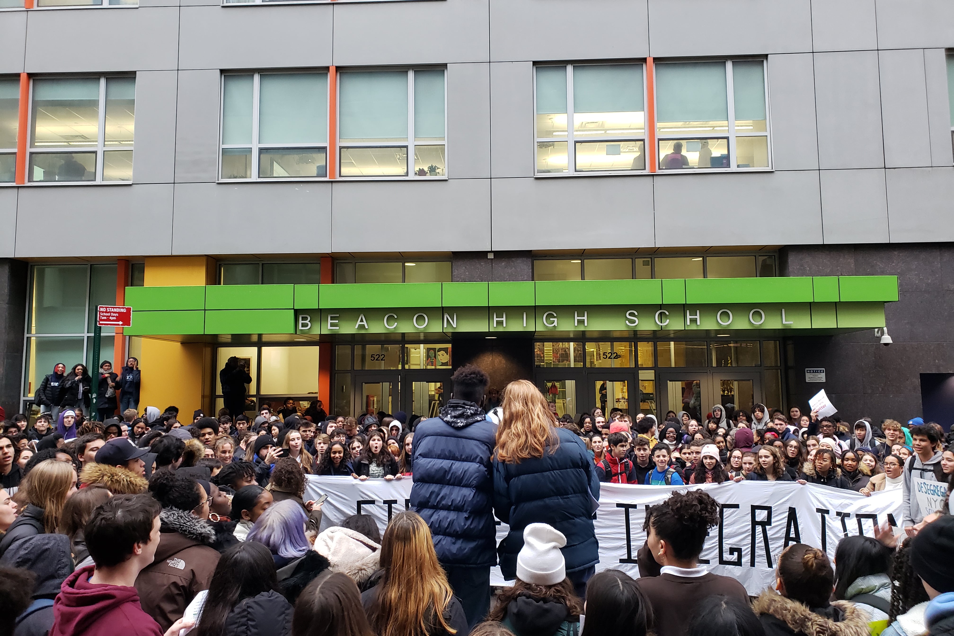 Hundreds of teens gather in front of a high school building.