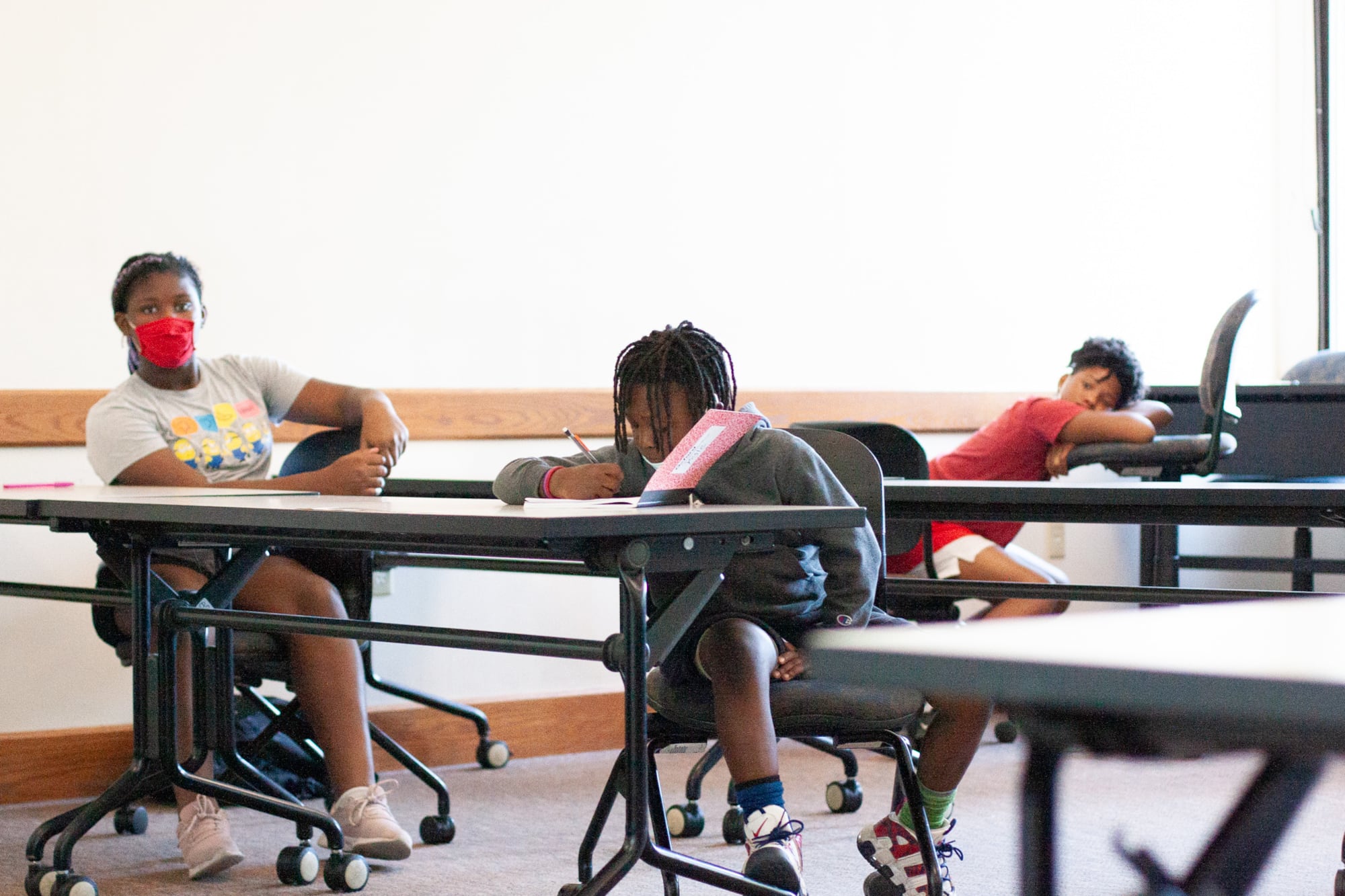 Three students sit at large desks in a classroom. The student in the center is taking notes in a red composition notebook, the student on the left is wearing a red mask and listening to a lecture as the student behind them leans on a raised chair and listens.