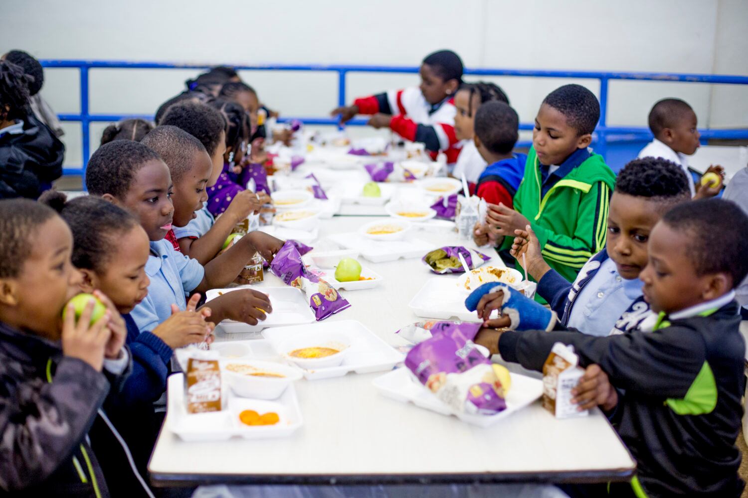 Young children sit at a long white table and eat lunch in a school cafeteria.