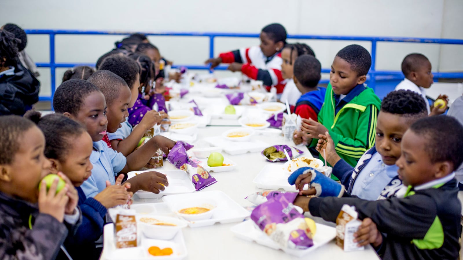 Young children sit at a long white table and eat lunch in a school cafeteria.
