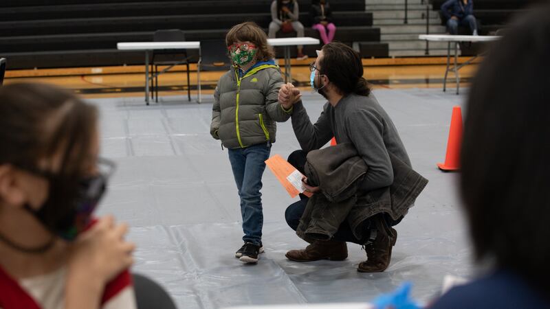 A young boy wearing a winter coat is comforted by an adult kneeling next to him before getting a vaccine shot inside a school gym.