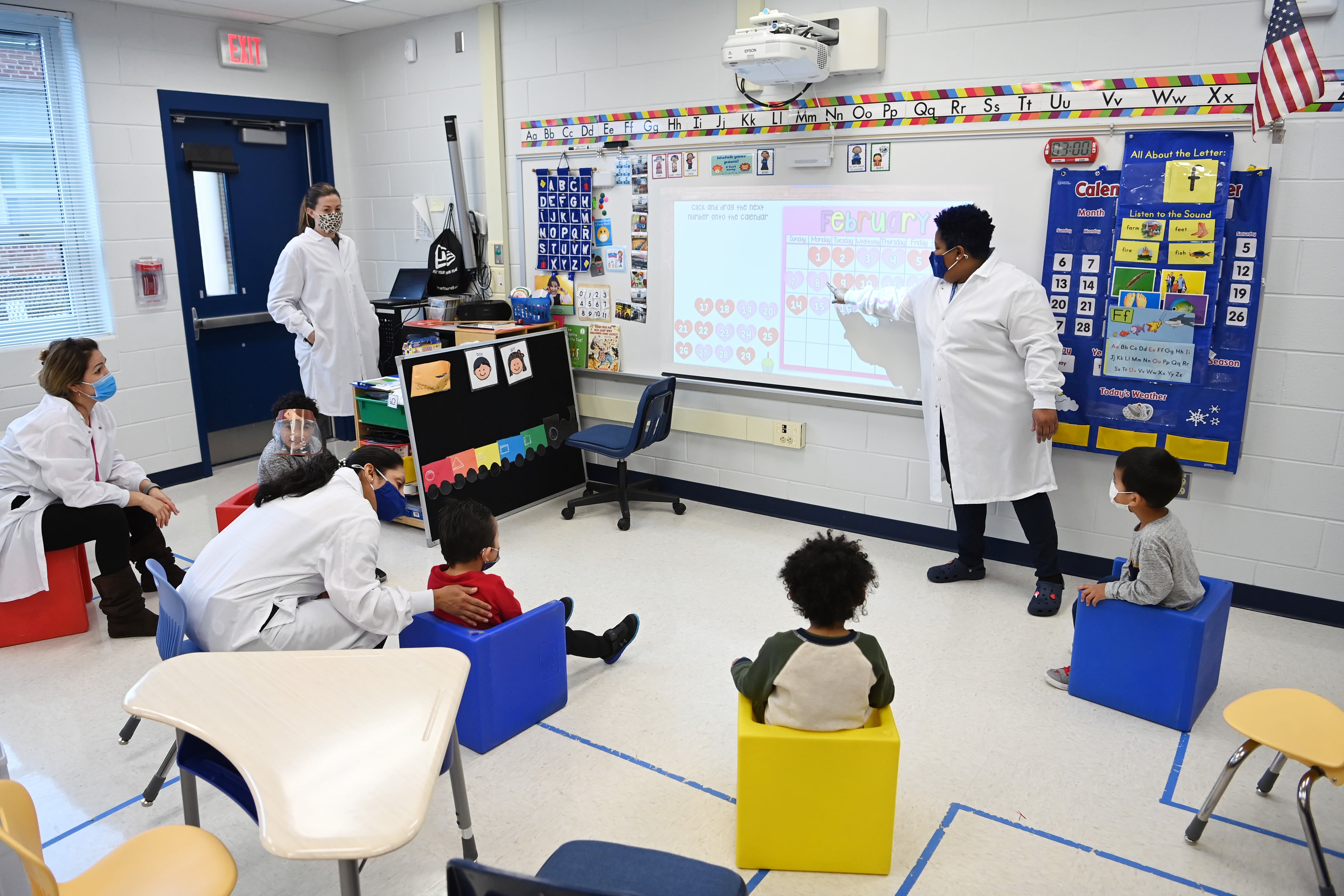 A teacher wearing a white lab coat and face mask points at a picture projected at the front of the class while students wearing face masks sit and watch.