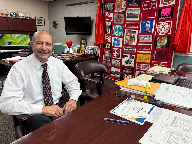 A photograph of a man in a white dress shirt and red tie sits at his desk in an office posing for a portrait.