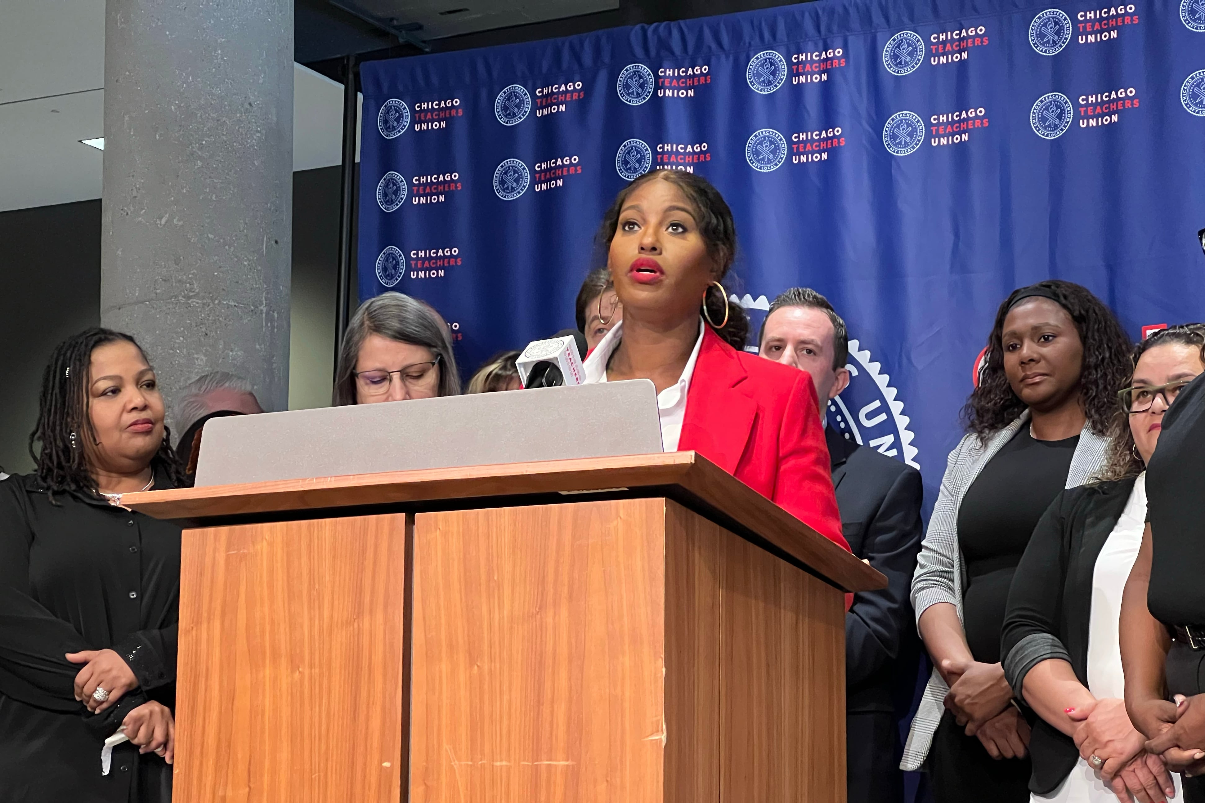 A Black woman wearing a red suit jacket speaks from behind a wooden podium and in front of a blue background an a group of people stand next to her.