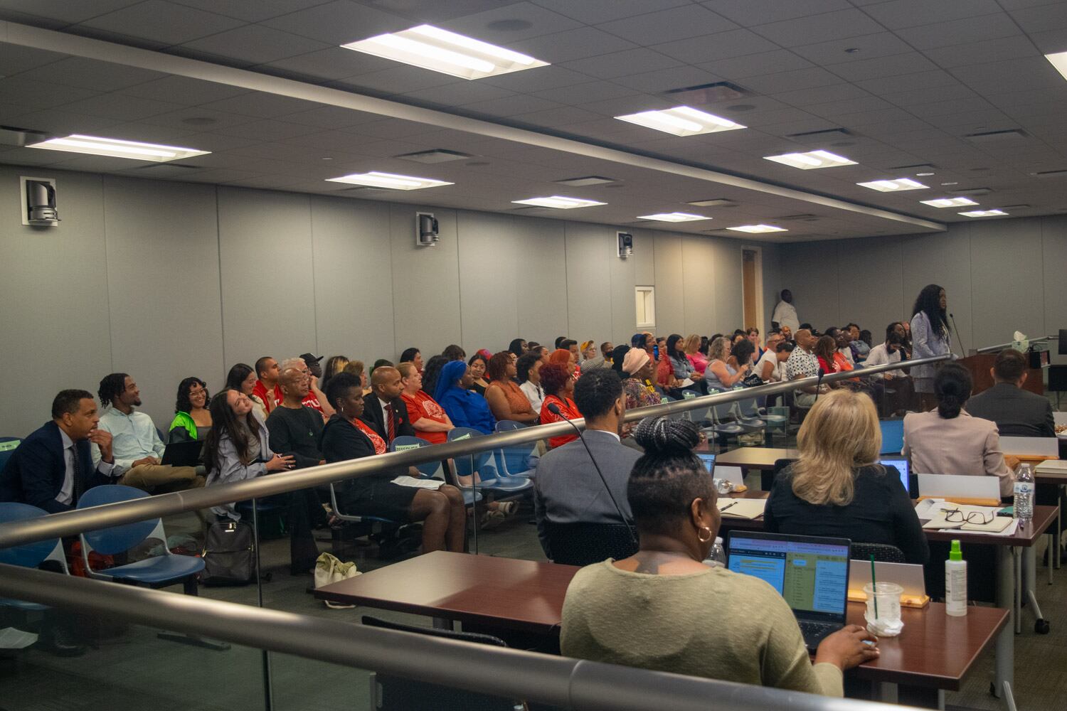 Nearly every seat in the board room is filled. At the podium is the Chicago Teachers Union president speaking. Behind a metal bar are Chicago Public Schools employees looking at their laptops.