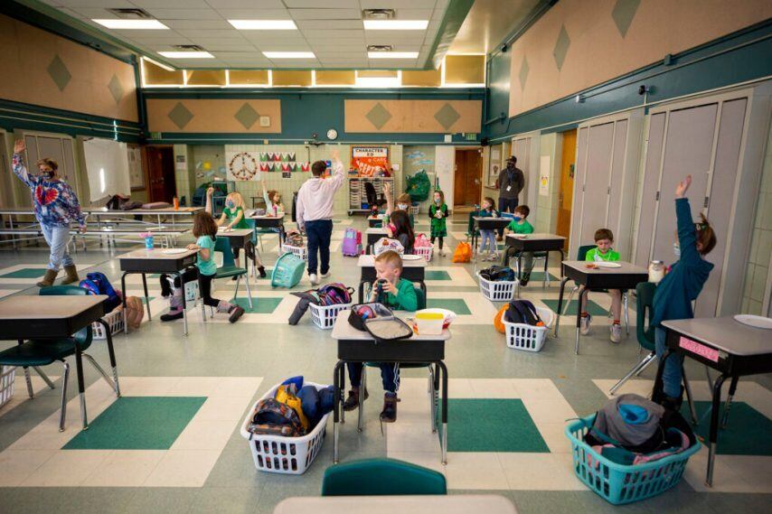 Ten students sit or stand at single desks, space out, in a classroom while three adults stand in different parts of the classroom, two with their arms up. Baskets holding backpacks sit next to the desks.