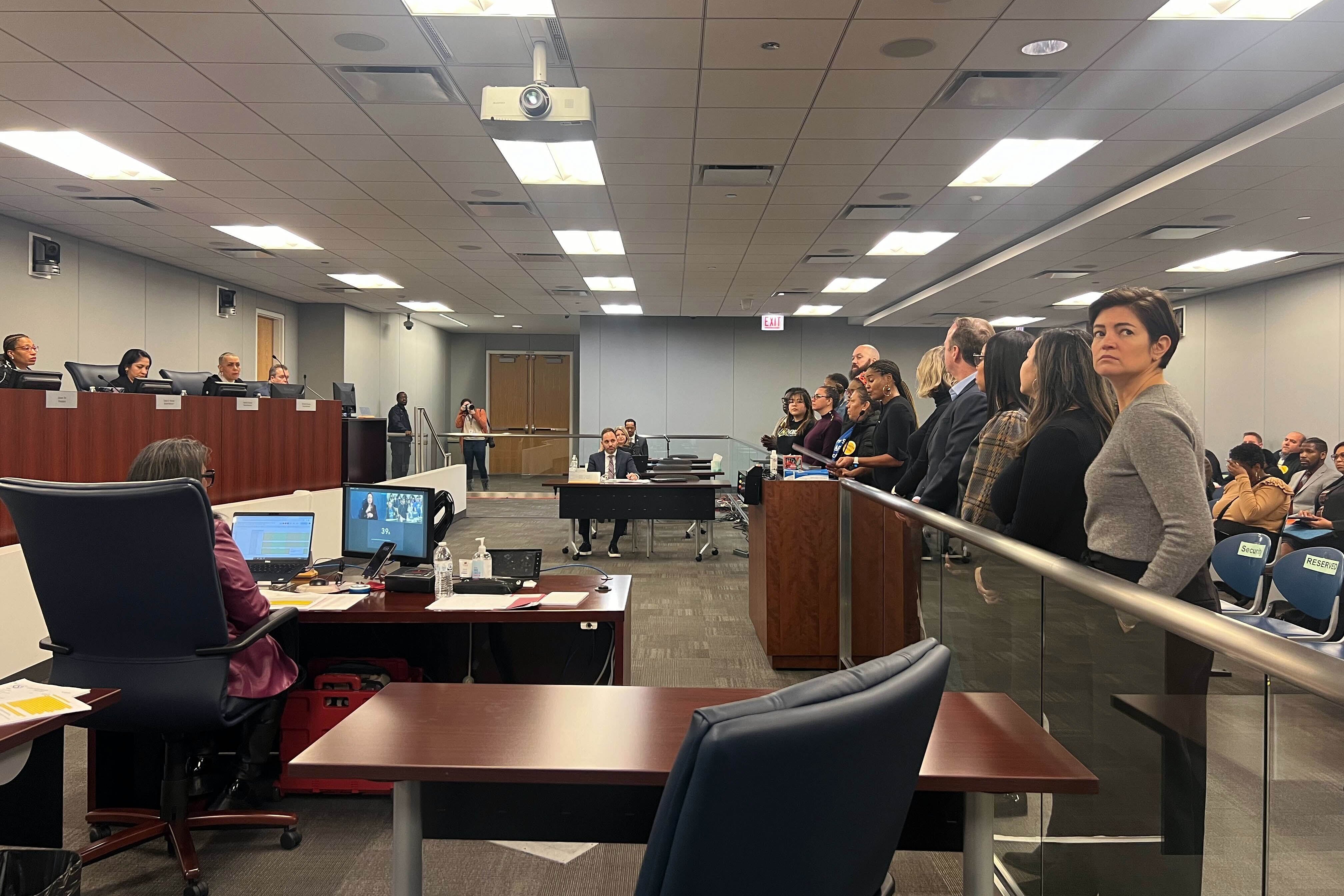 People sit behind a large wooden desk that lines the wall on the left while people stand behind a bar in a room filled with wooden desks and chairs.