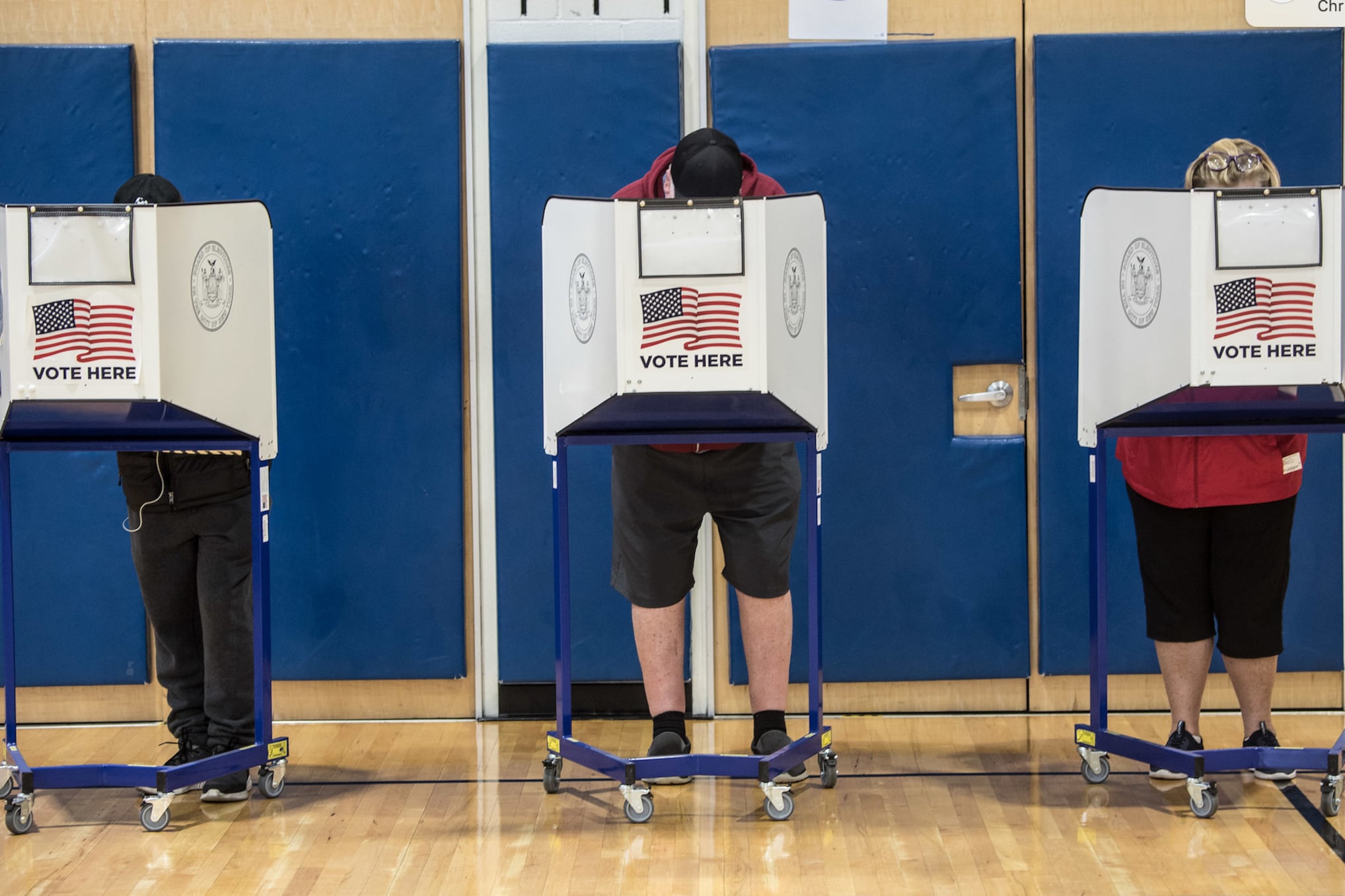Three people stand behind voting partitions against a blue divider.