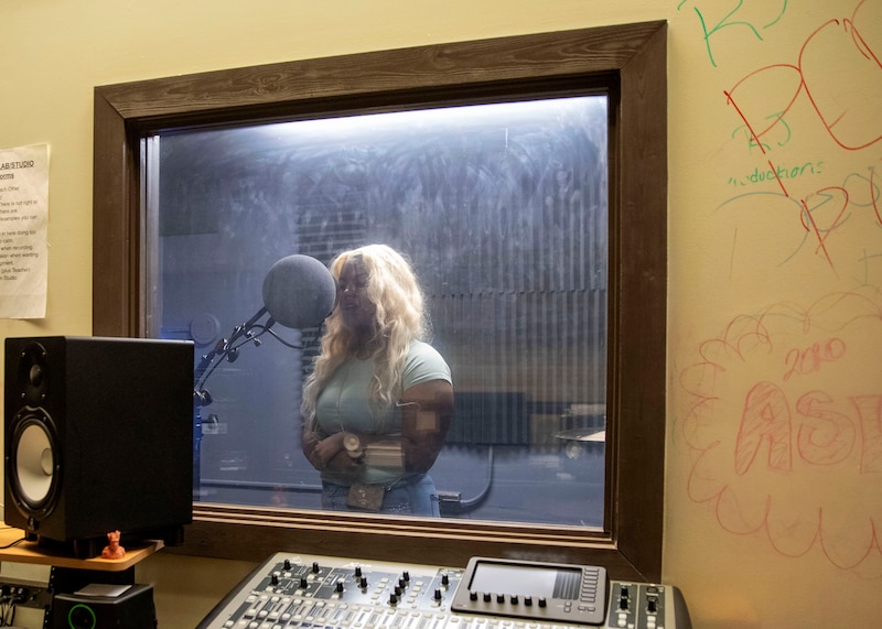 A photograph of a Black high school student singing in a recording studio. She has long blonde hair.