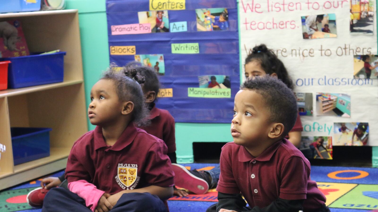 Toddlers wearing red collared shirts sit on a colorful rug in a classroom.