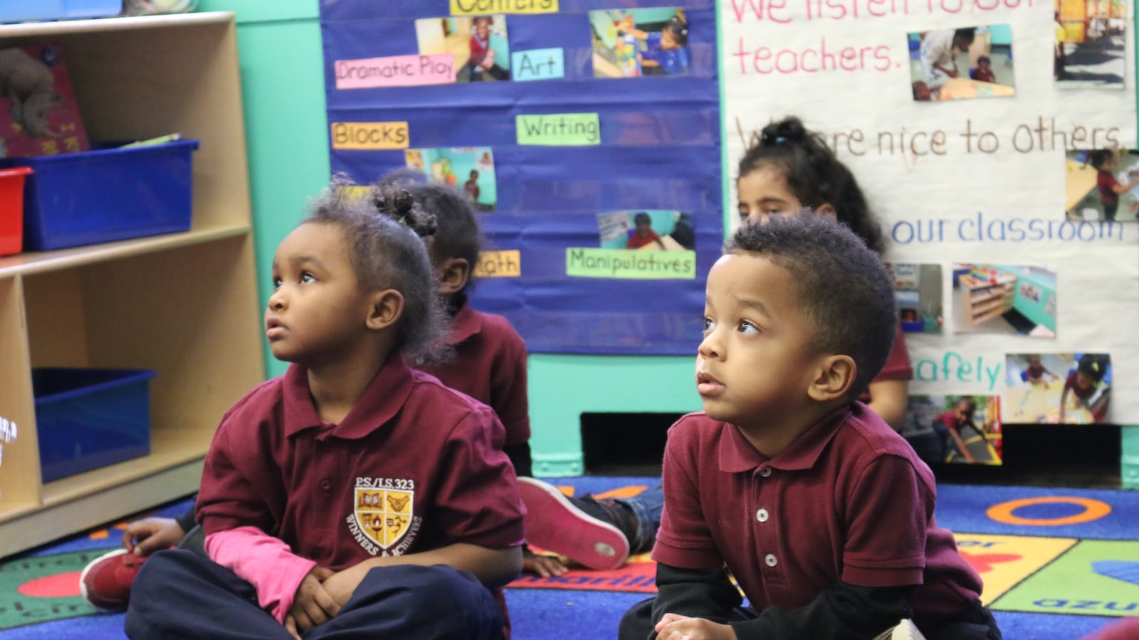 Toddlers wearing red collared shirts sit on a colorful rug in a classroom.