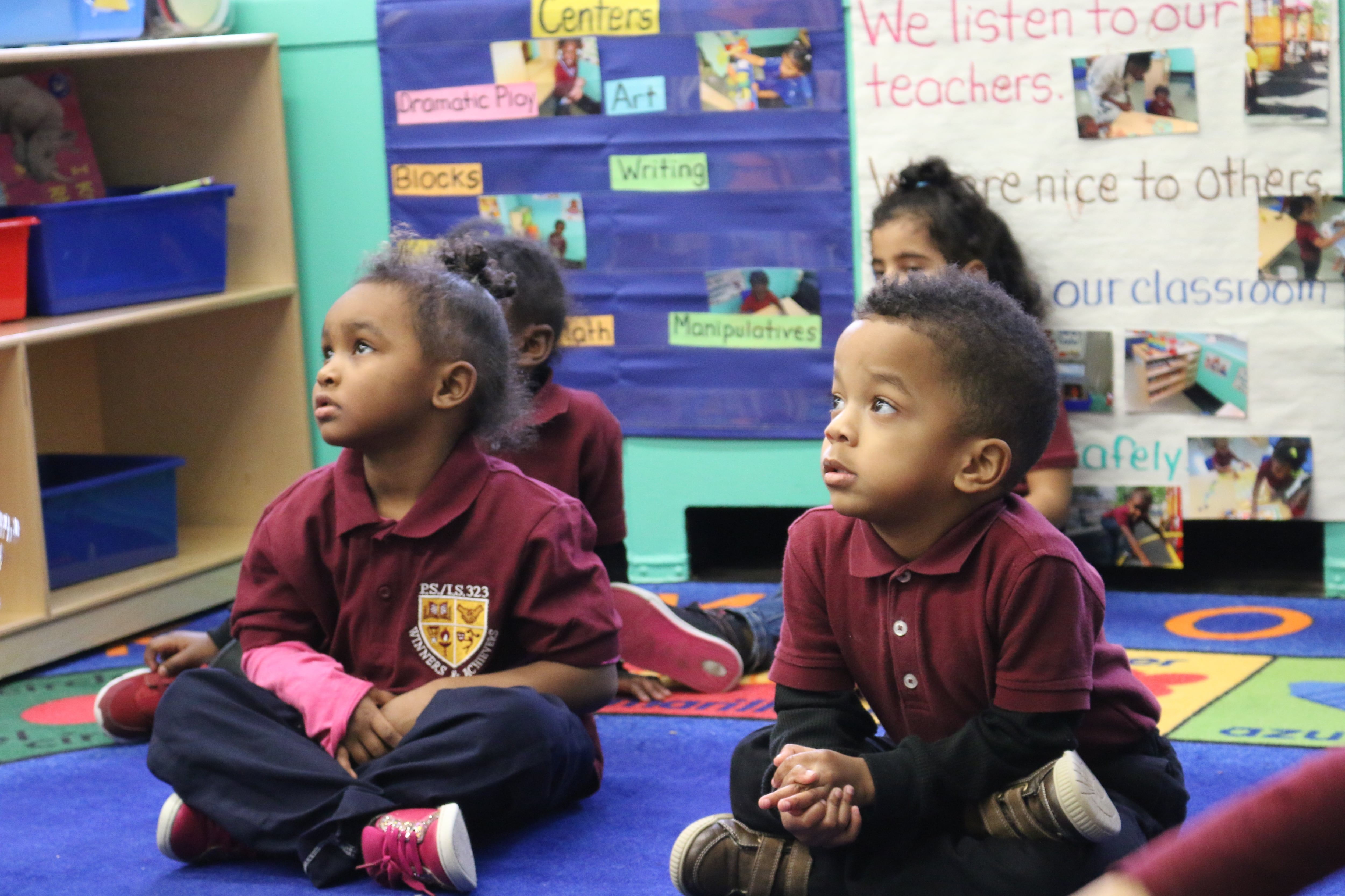 Toddlers wearing red collared shirts sit on a colorful rug in a classroom.