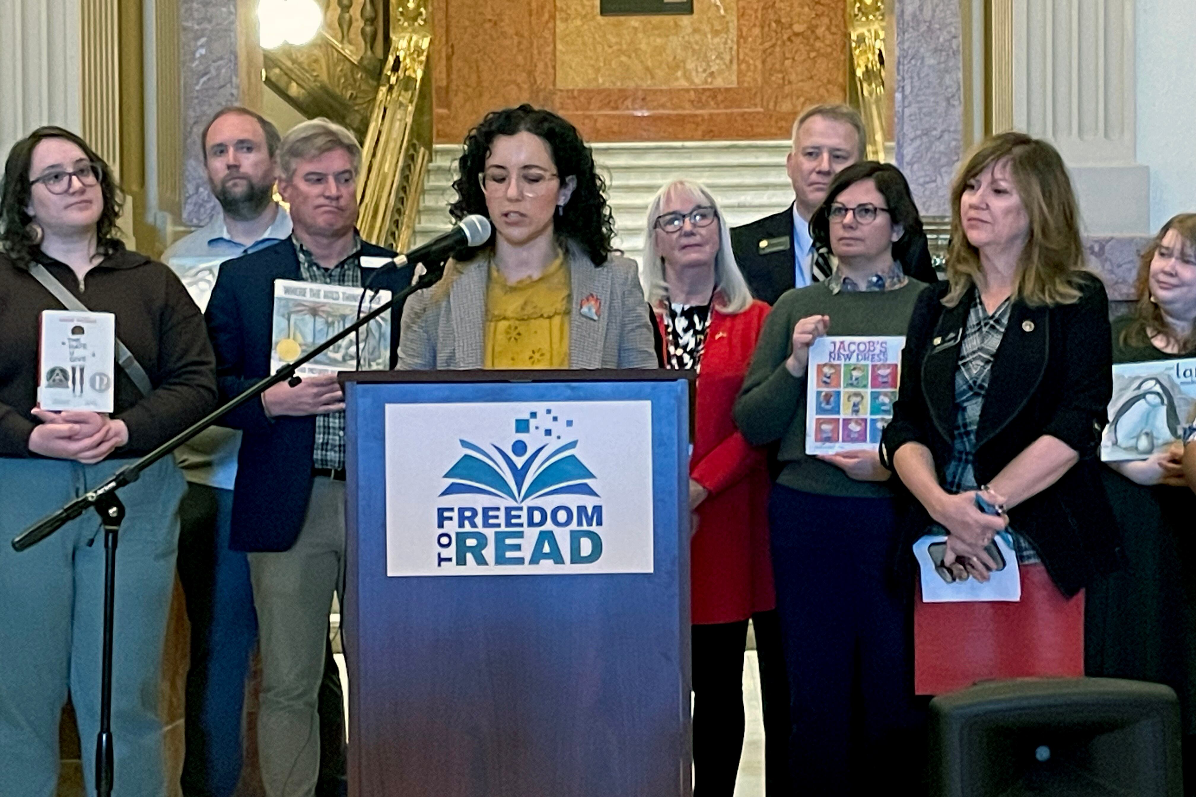 A woman with short dark hair and wearing a suit speaks from a wooden podium with a group of people standing behind her.