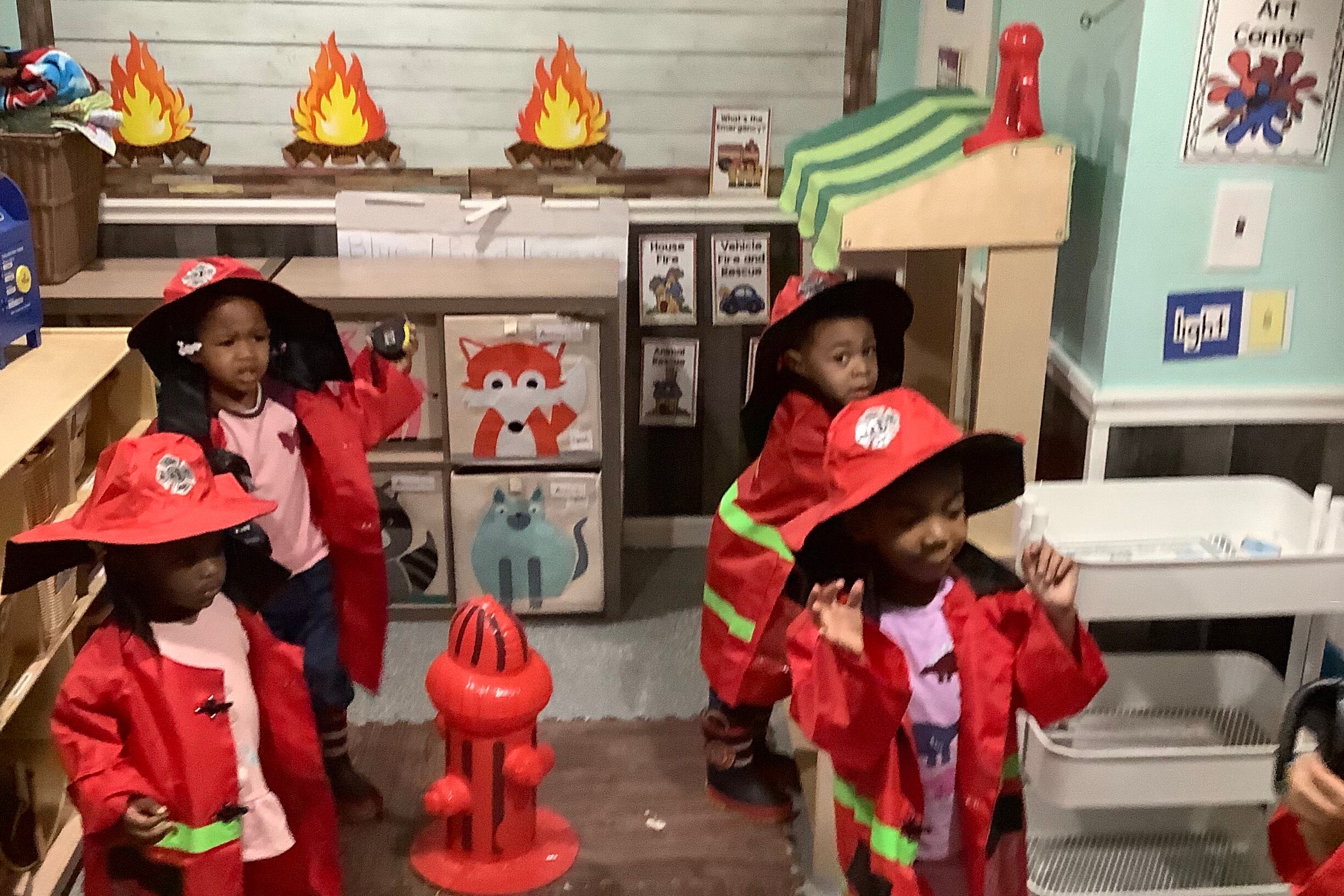 Children wear pretend red fire outfits and helmets next to a pretend red fire hydrant in a preschool classroom