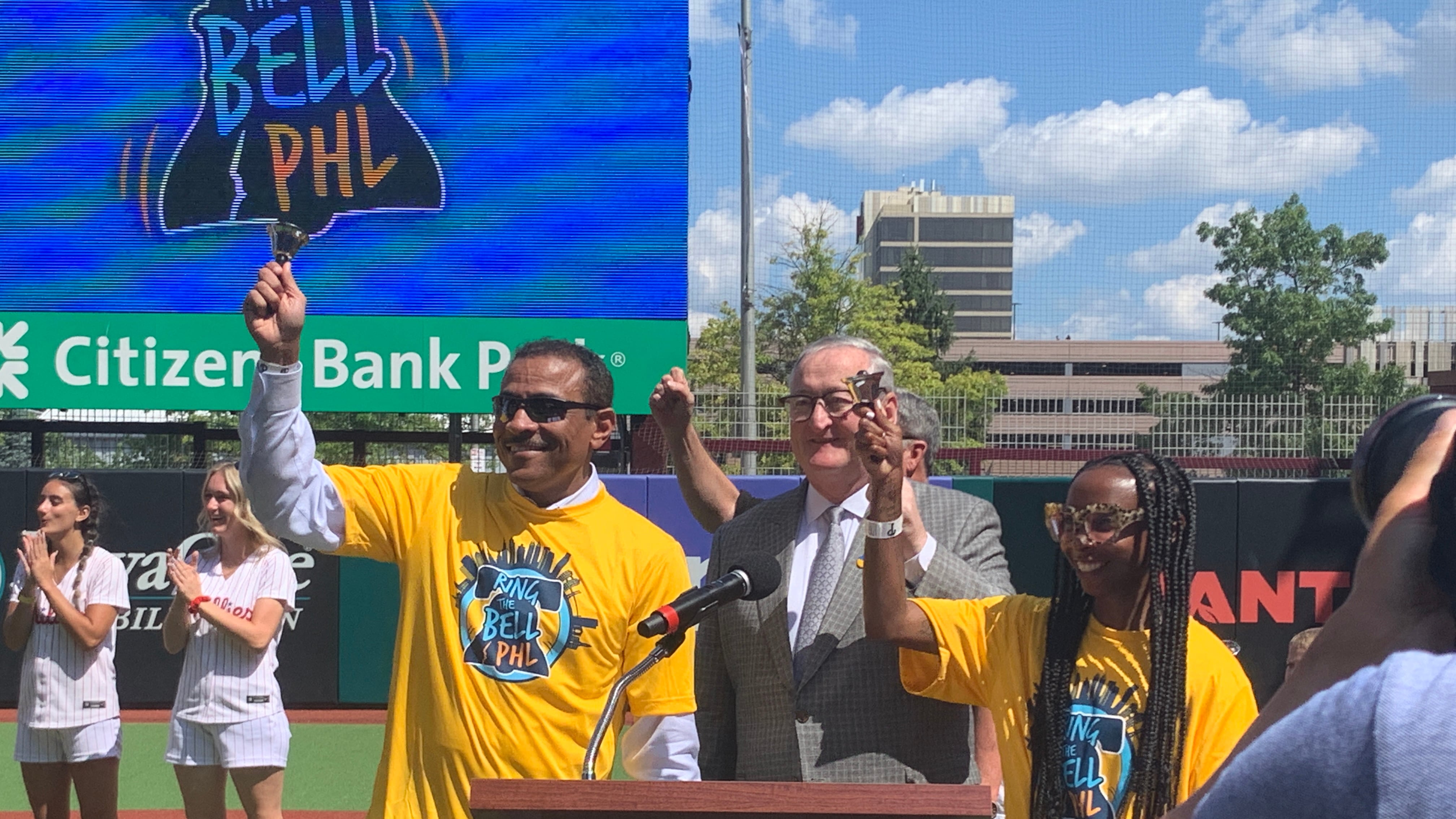 A man in a yellow T-shirt and white long sleeve shirt raises his right hand and rings a bell with it while standing behind a podium on a baseball field. A man in a suit and a woman in a yellow T-shirt also stand behind the podium.