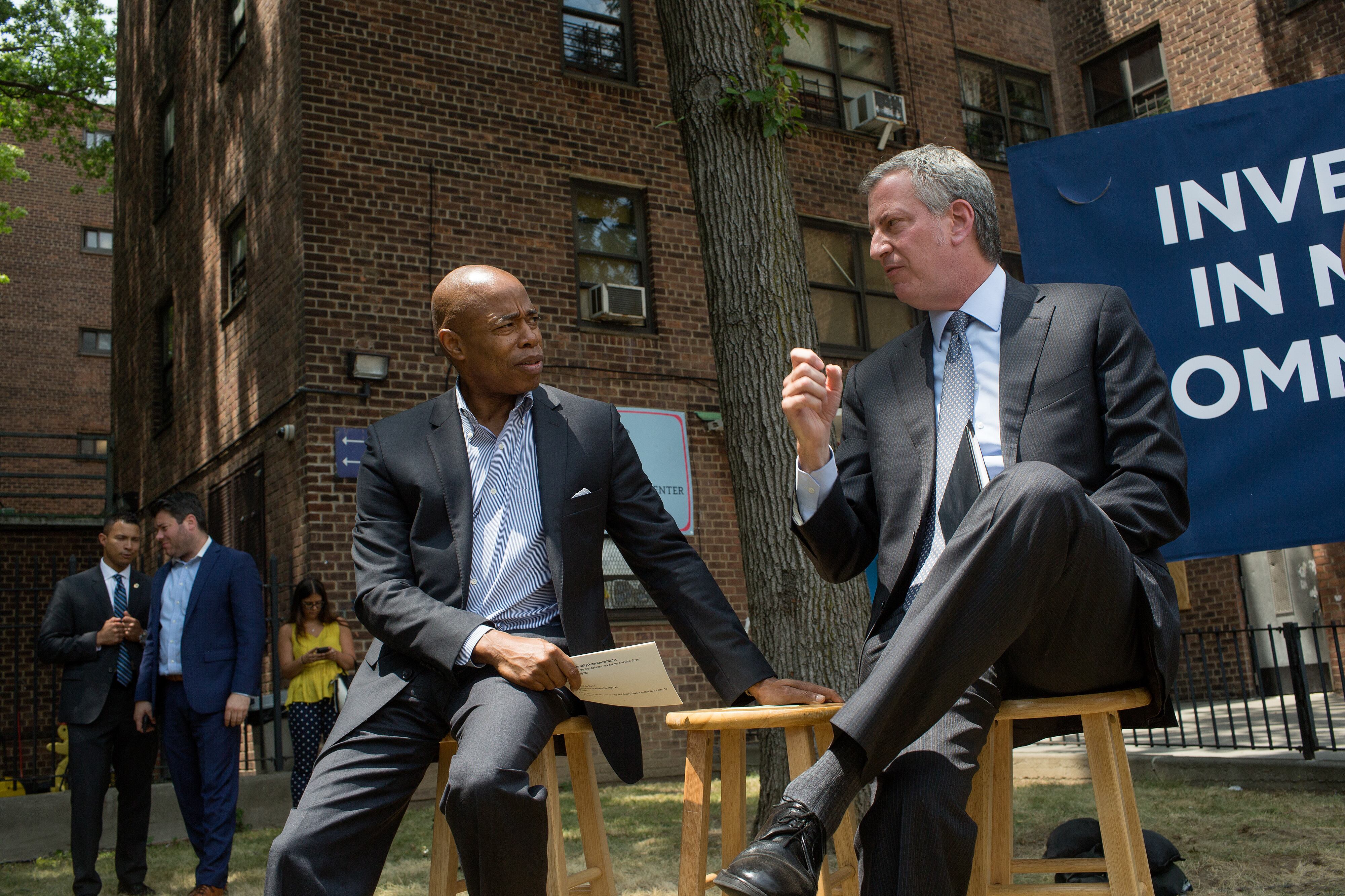 Eric Adams and Bill de Blasio speak together while sitting on wooden stools in front of a large brick building.