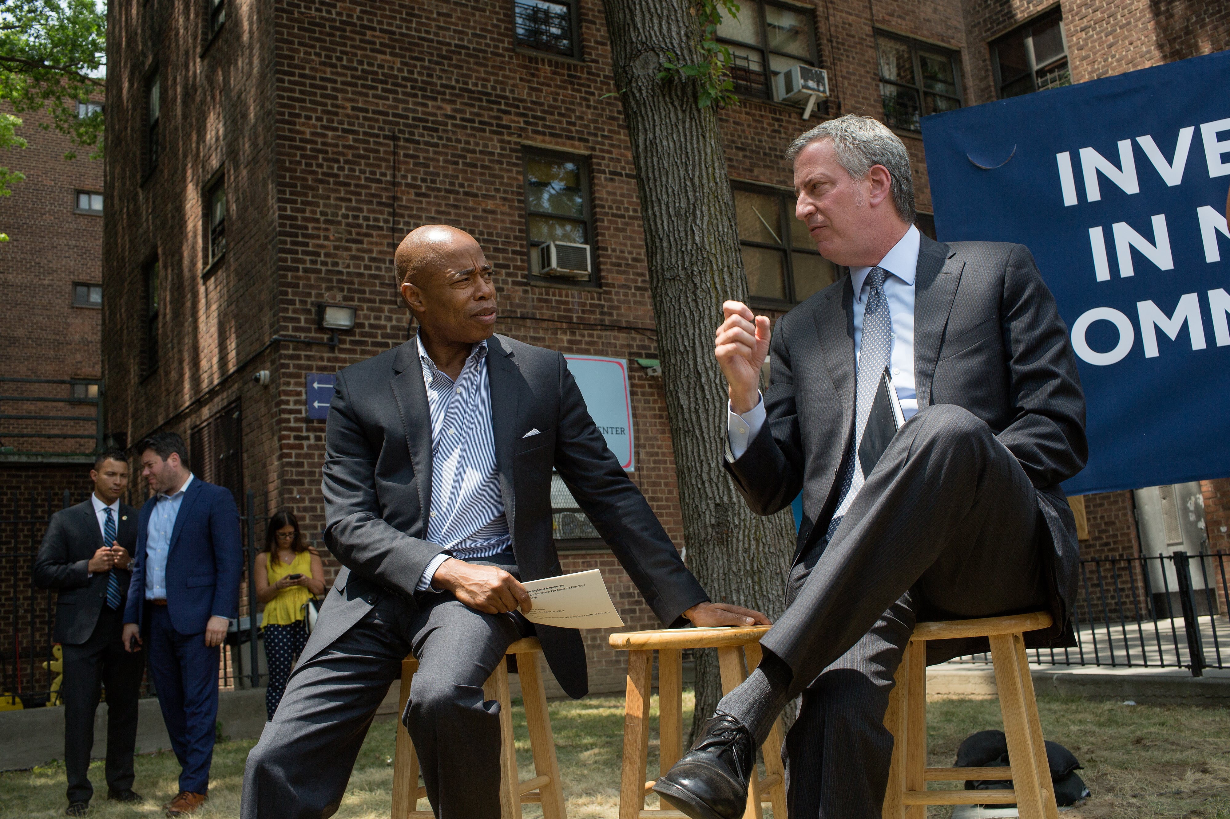 Eric Adams and Bill de Blasio speak together while sitting on wooden stools in front of a large brick building.