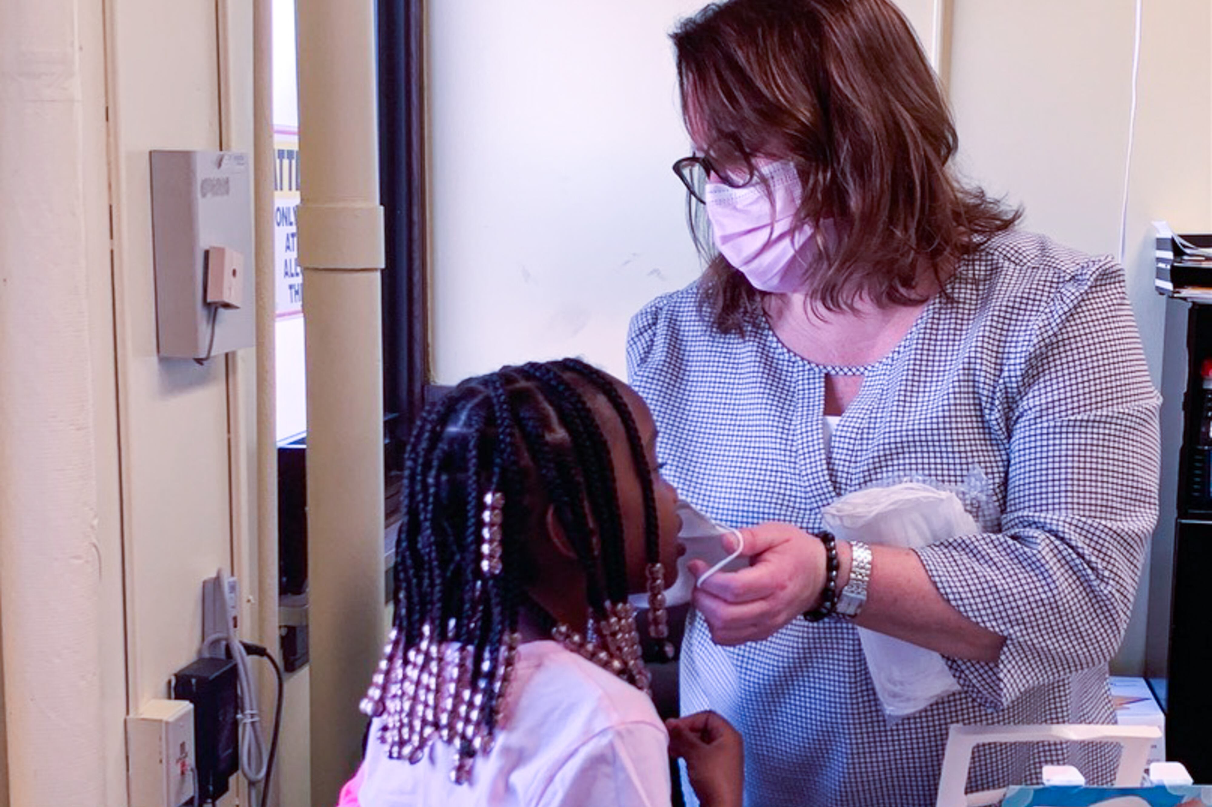 In an office, woman helps a little girl put on a mask.