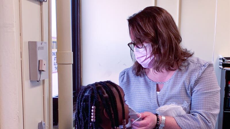 In an office, woman helps a little girl put on a mask.