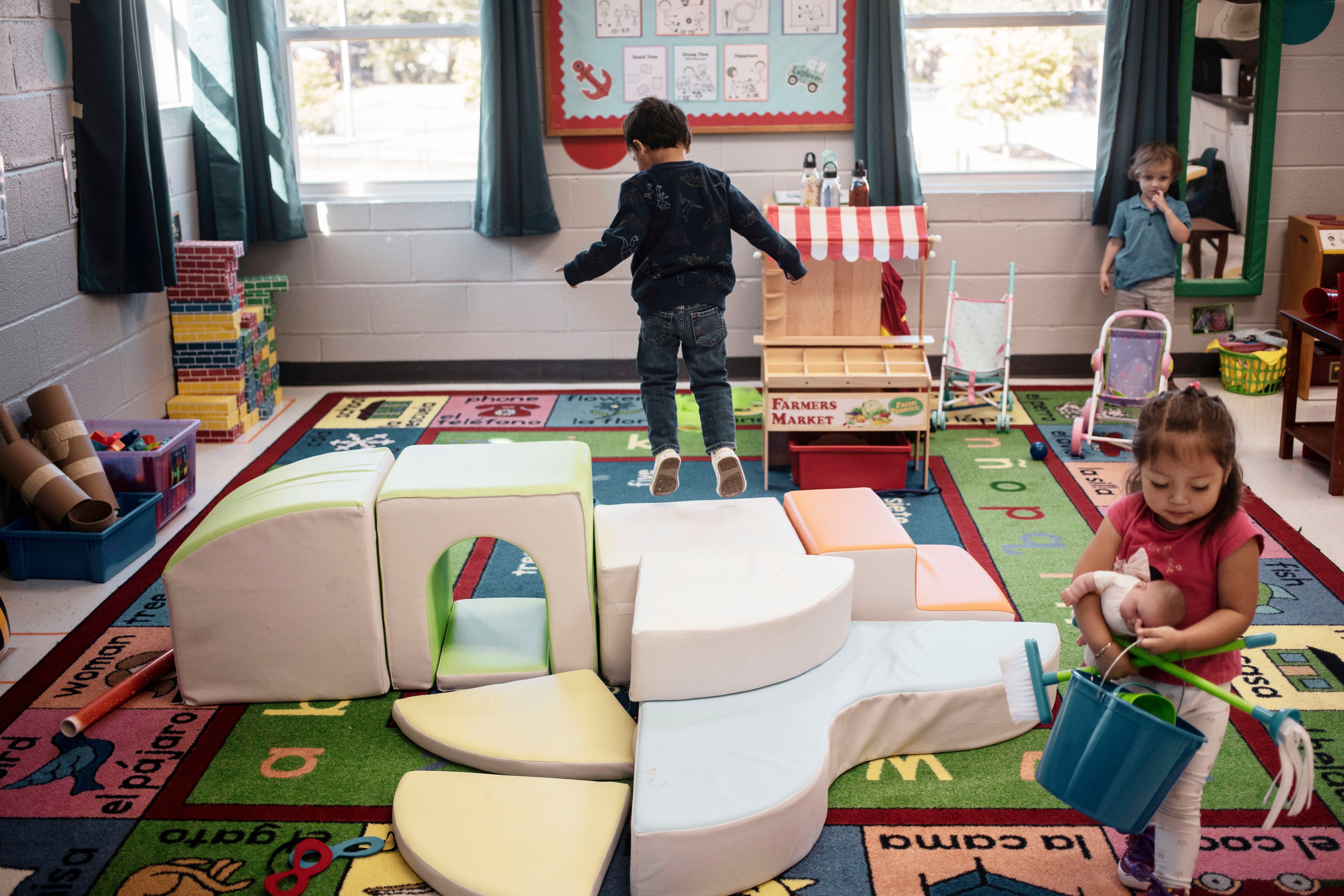 A little boy jumps off of a plush set of steps. A little girl walks with a doll and a bucket of toys in the foreground as another boy stands near a window in the back of the classroom.