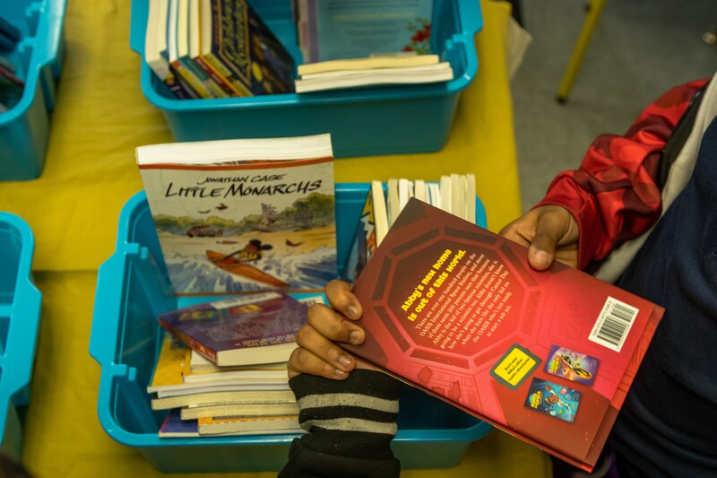 A turquoise basket full of books sits on a classroom desk. Hands of a student holding one of the books is shown.