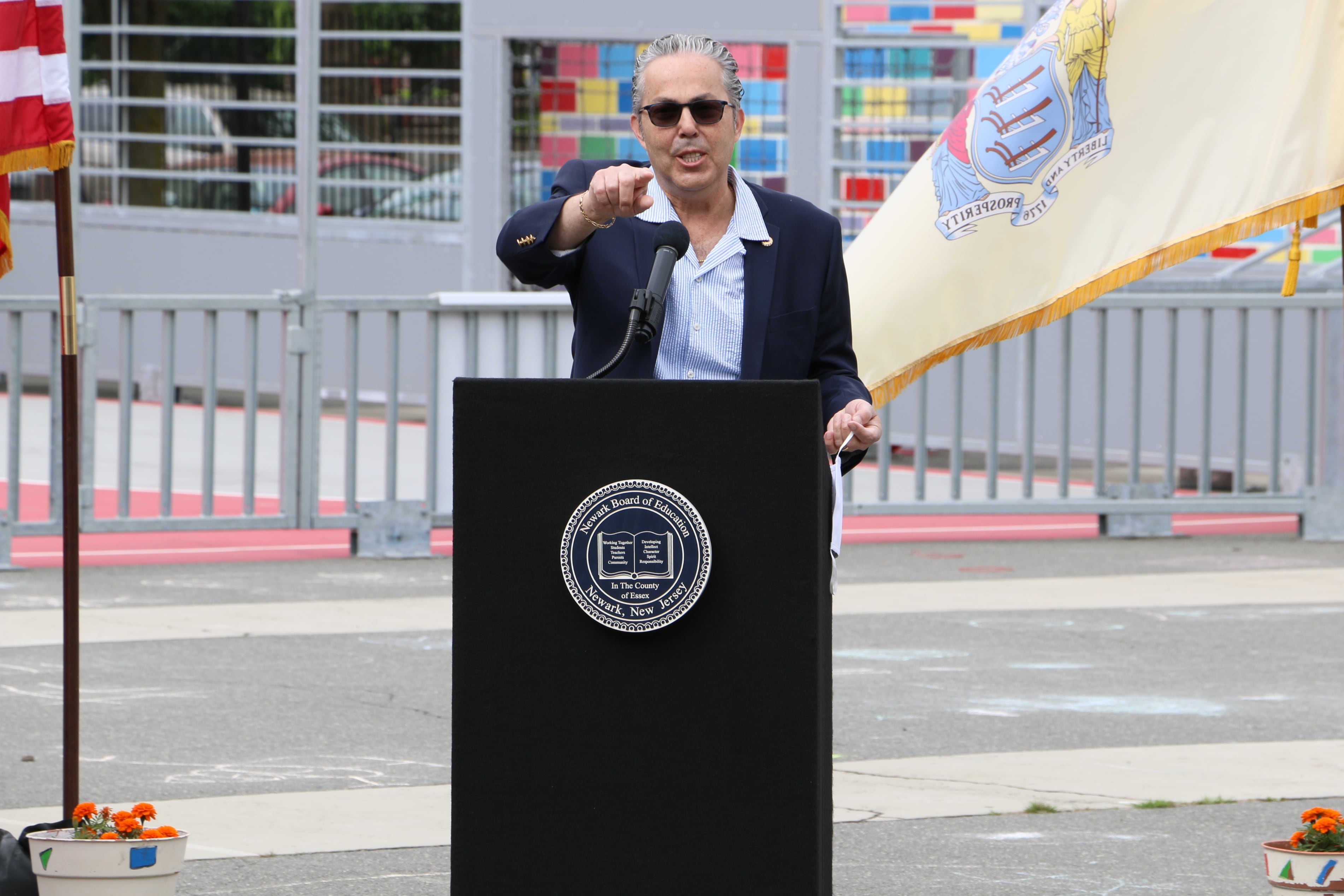 Man wearing sunglasses and a blue button-up shirt with a black blazer stands at a podium pointing in the direction of the camera.