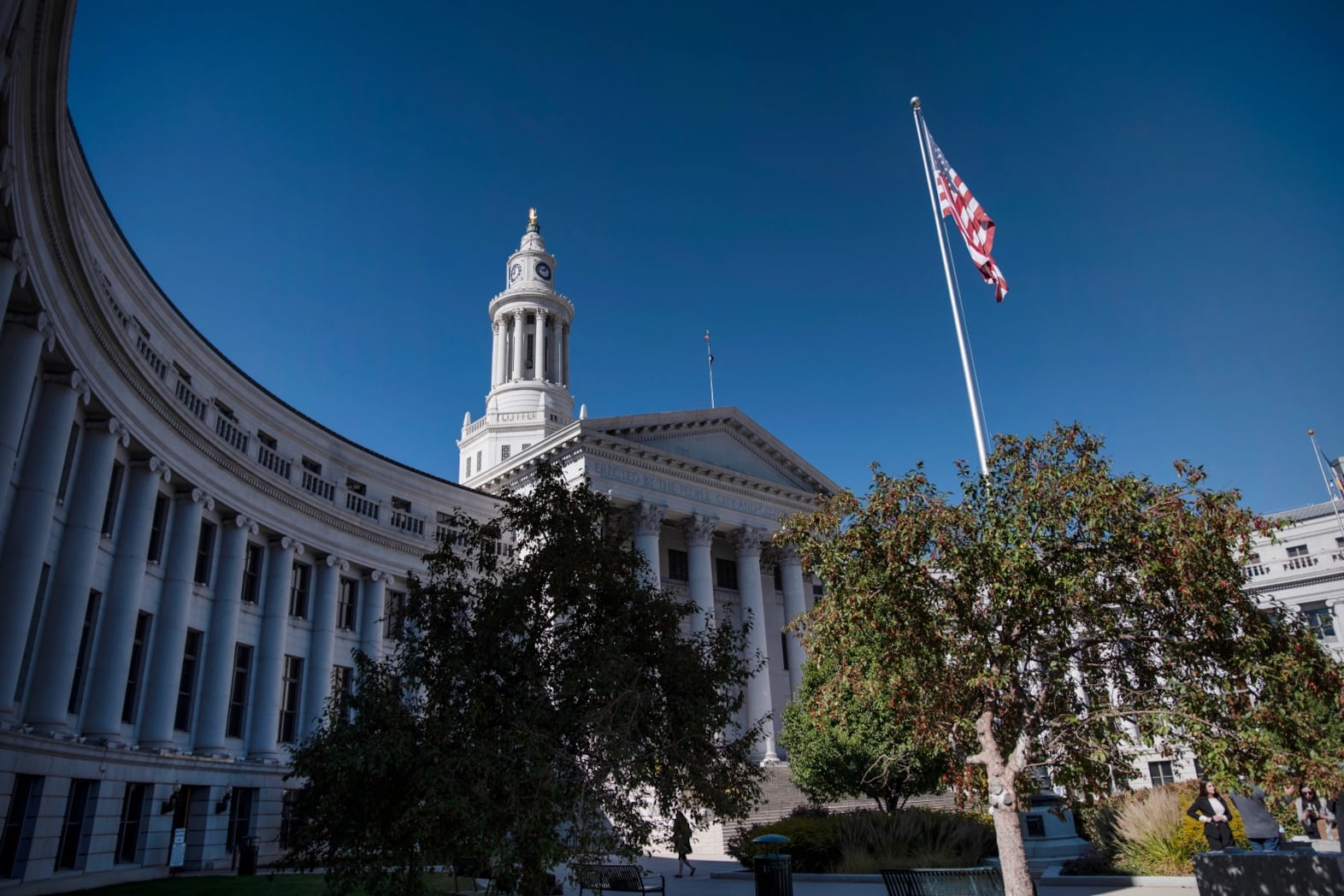 A white building with columns stands against a bright blue sky. An American flag is visible.