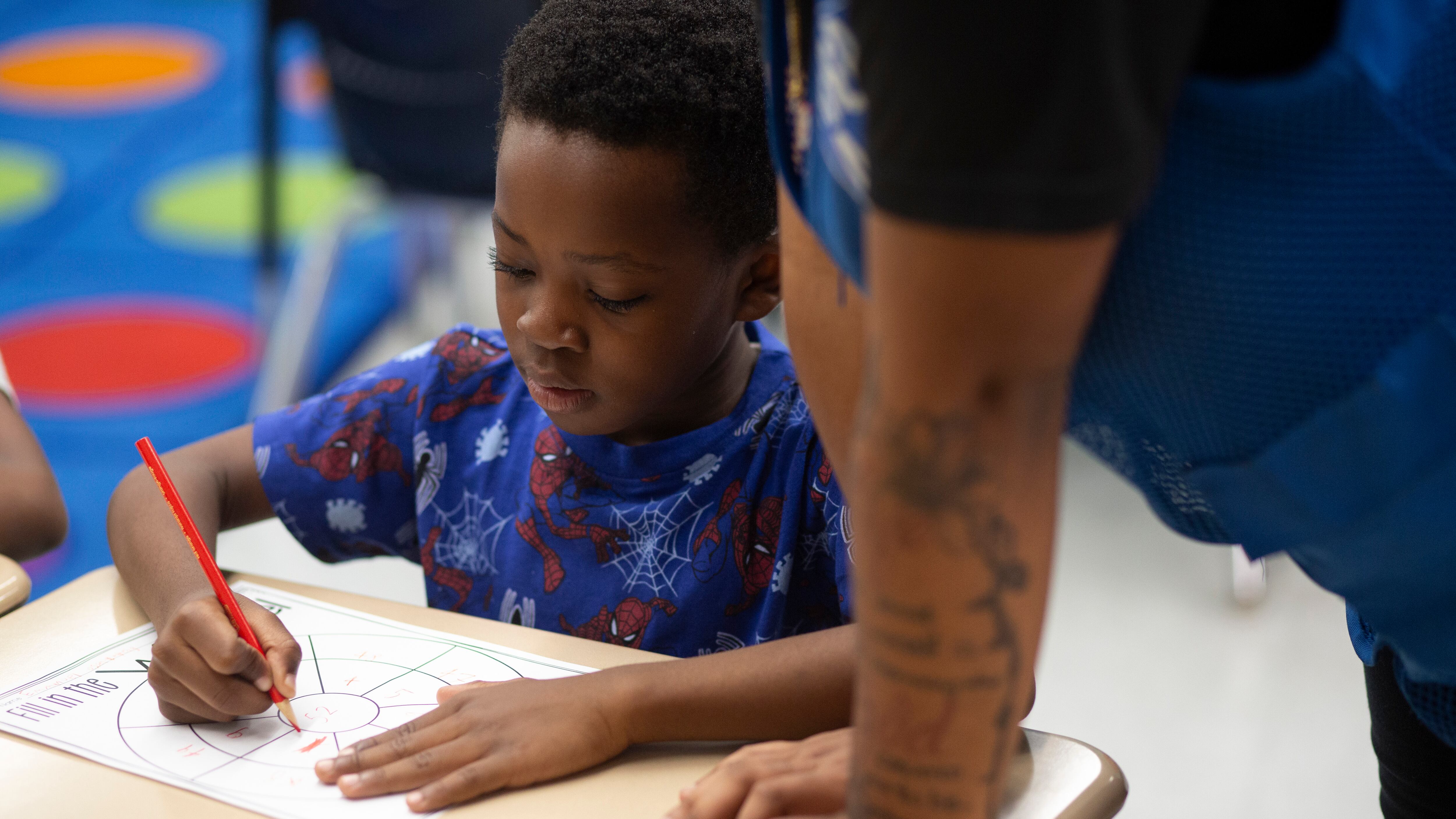 A boy wearing a blue shirt sits at a desk as a teacher’s aide helps him with his math work.