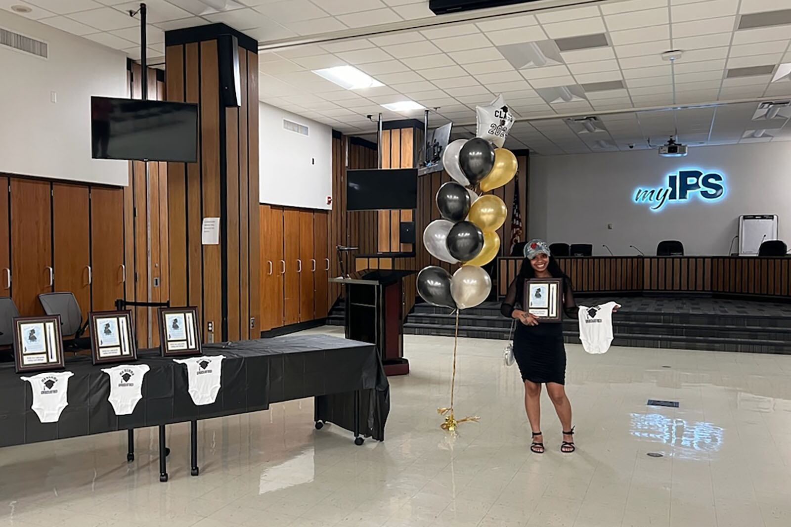 A graduating student stands to the left of a long table with a black table cloth with onesies and frames and graduation balloons floating in the middle. An illuminated IPS sign in the background.