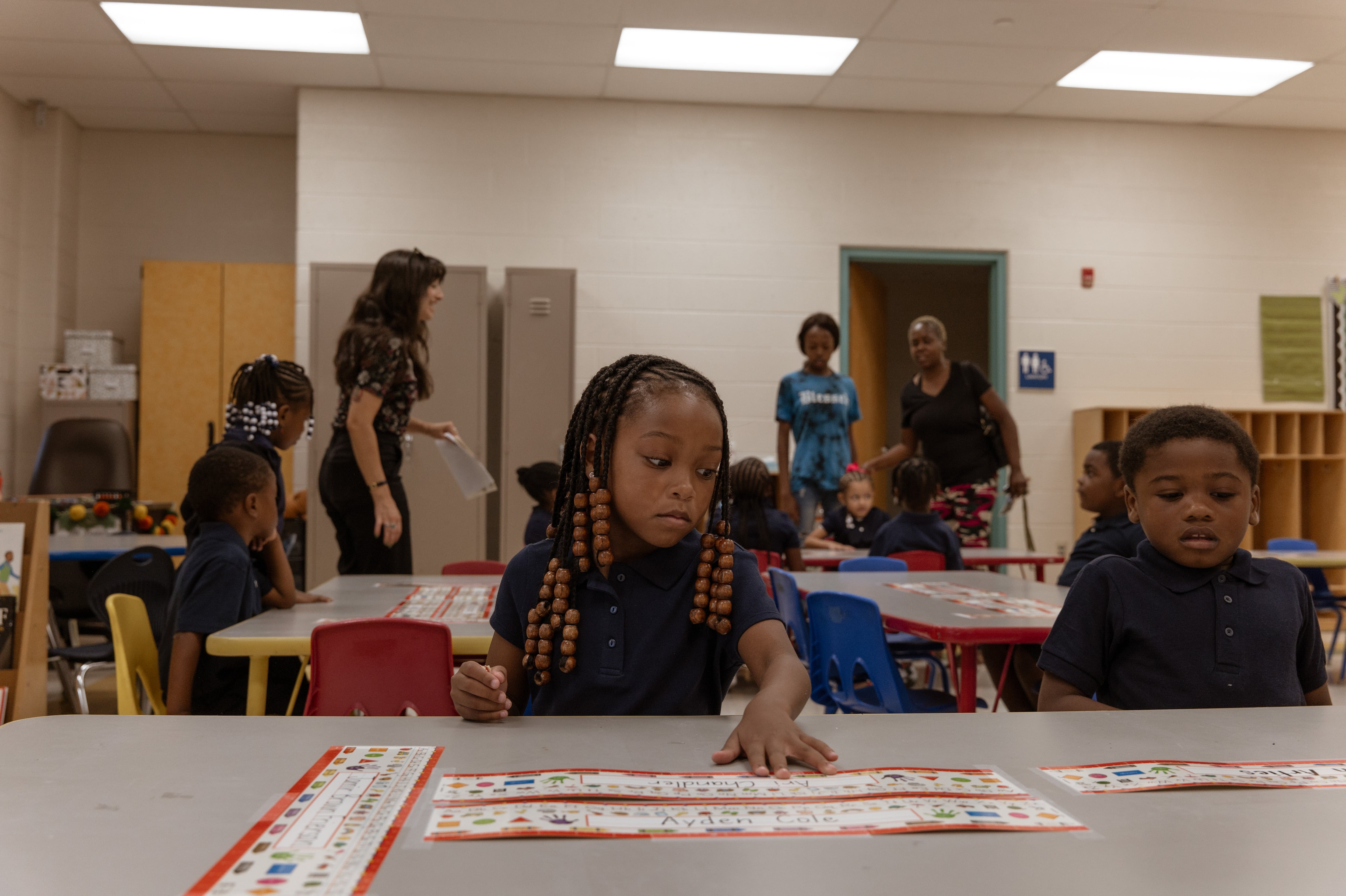 A girl with braids and a dark shirts puts her left hand on a table and looks to her left as a boy to her left in a dark shirt looks down at the table as people stand in the background.