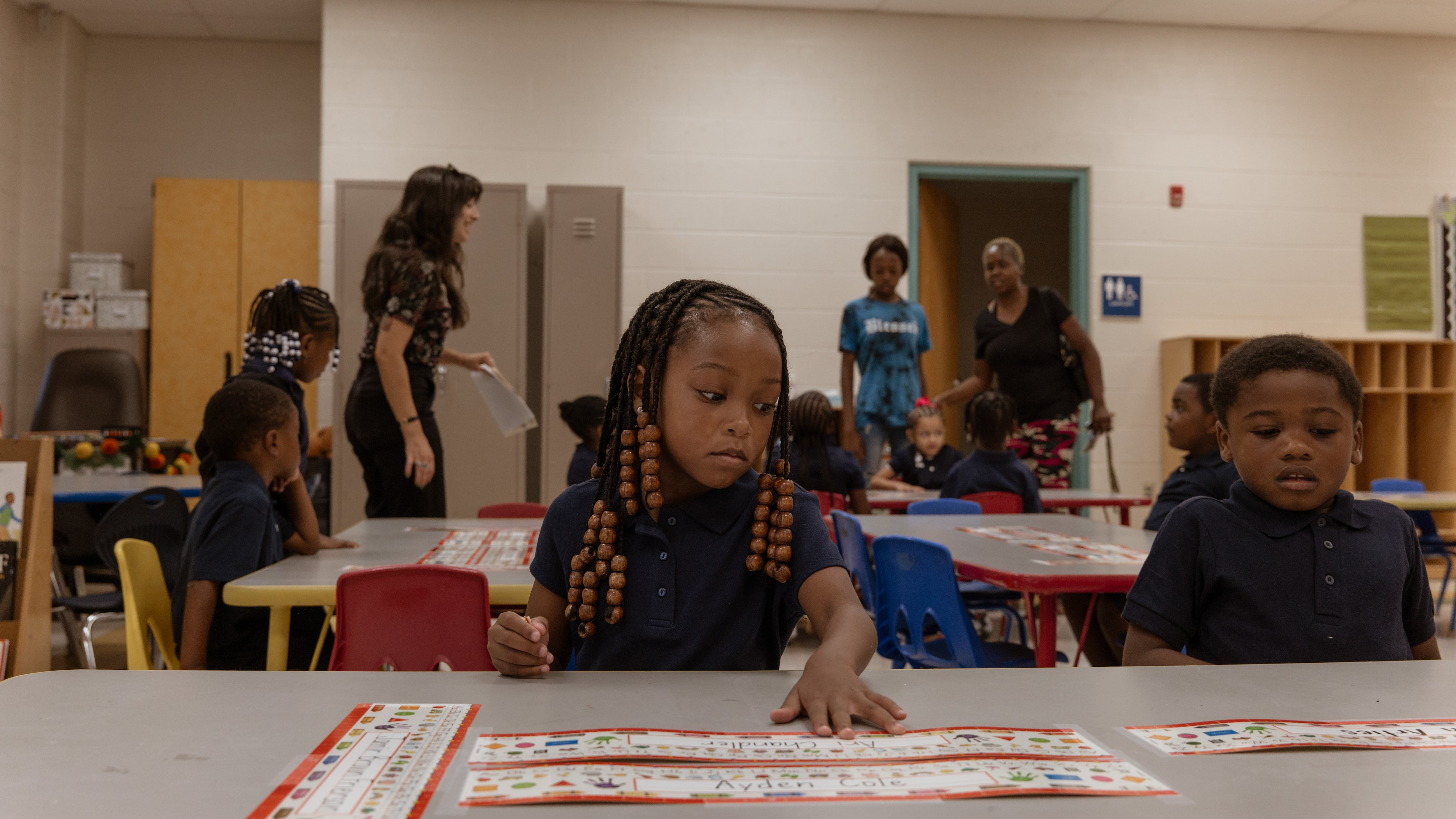 A girl with braids and a dark shirts puts her left hand on a table and looks to her left as a boy to her left in a dark shirt looks down at the table as people stand in the background. 