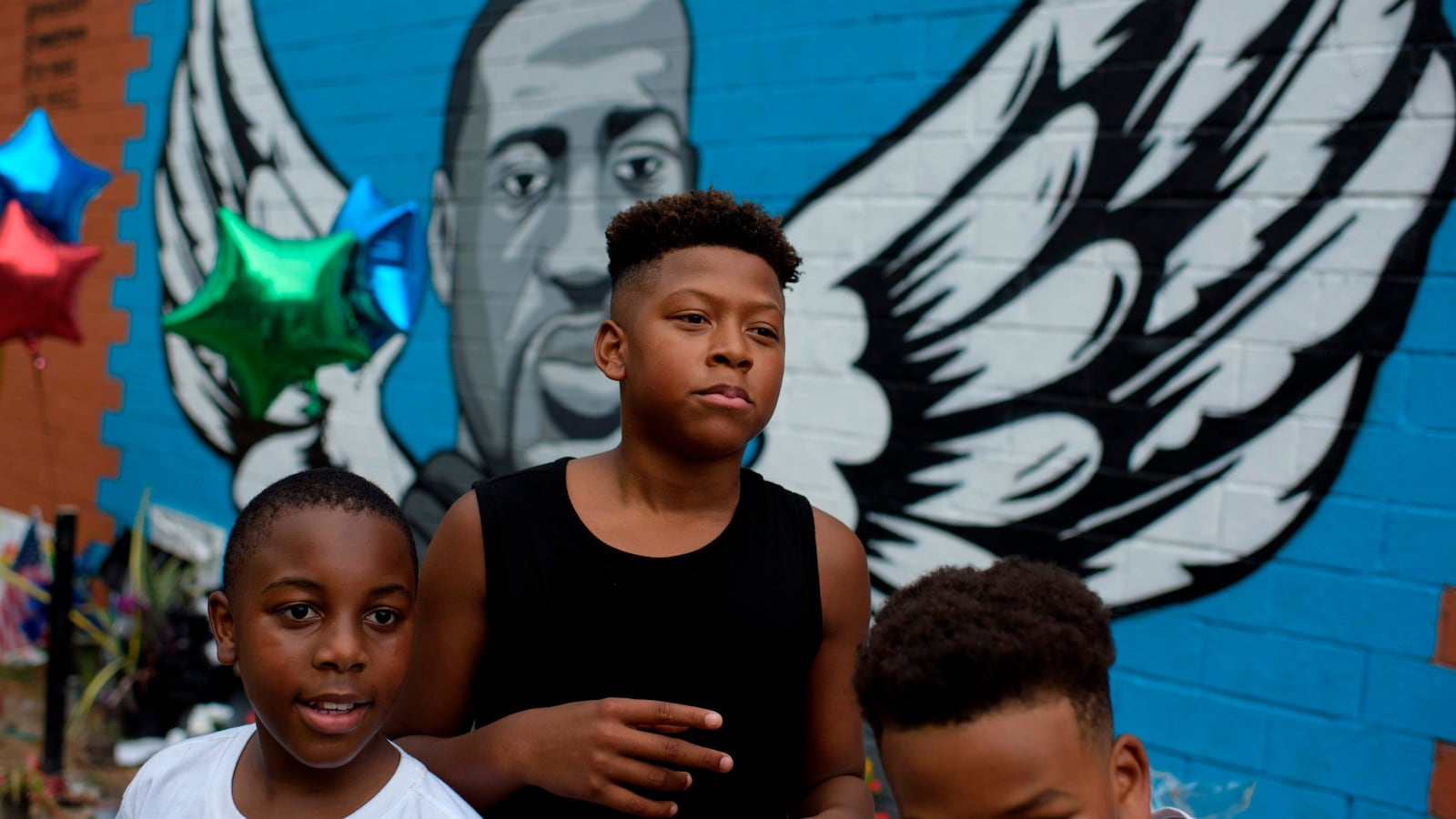 Three boys, one in a black tank top and two wearing white t-shirts, stand in front of a blue mural in honor of George Floyd. The mural has Floyd’s portrait with angel wings on either side.