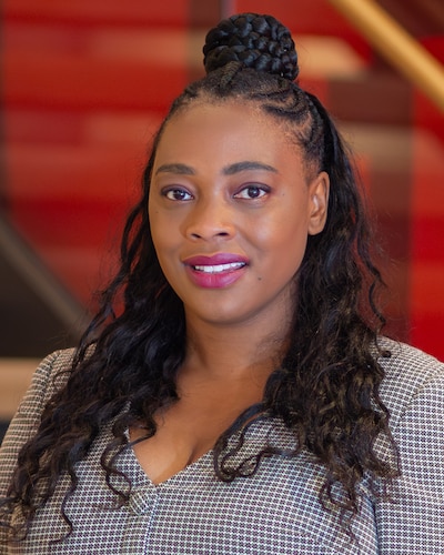 A photograph of a Black woman smiling for a portrait with long dark hair and a bun and wearing a blouse and standing in front of a red background.