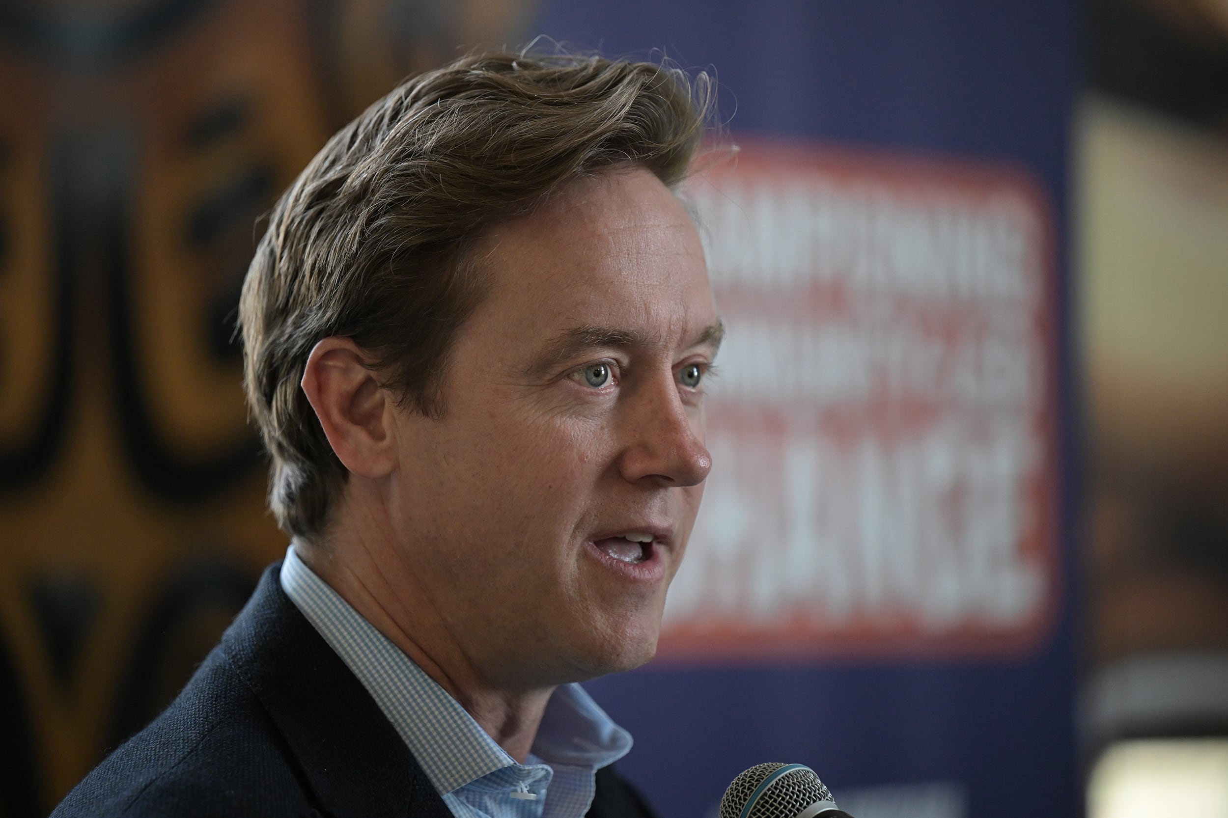 A photograph of a white man in a suit speaking in a room.