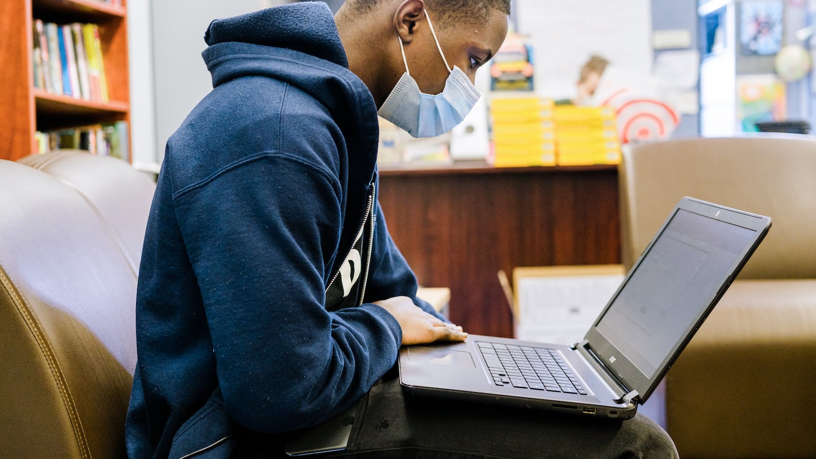 A teenager male in a blue sweatshirt is looking at a computer in a classroom. He is wearing a mask.
