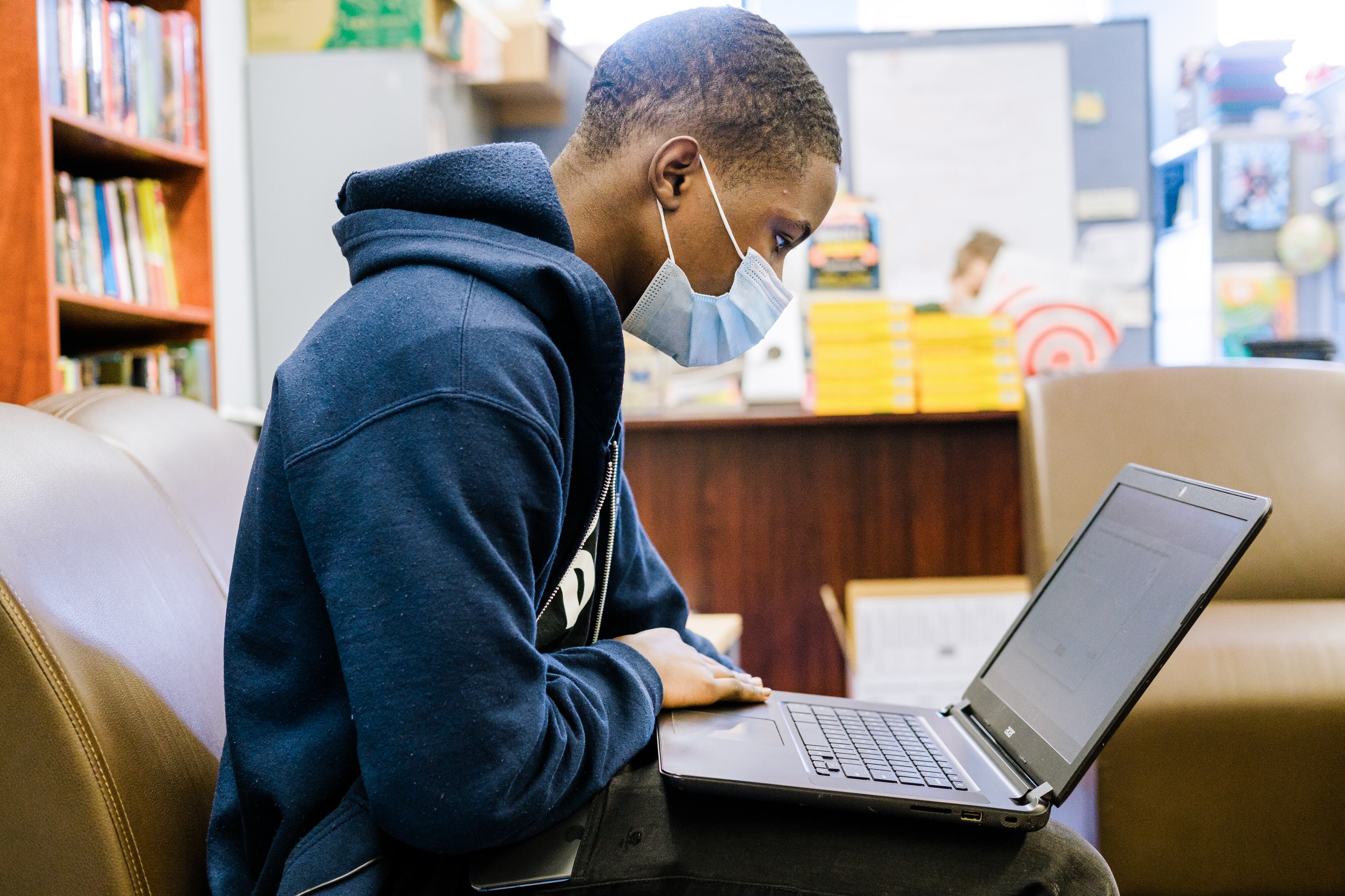 A teenager male in a blue sweatshirt is looking at a computer in a classroom. He is wearing a mask.