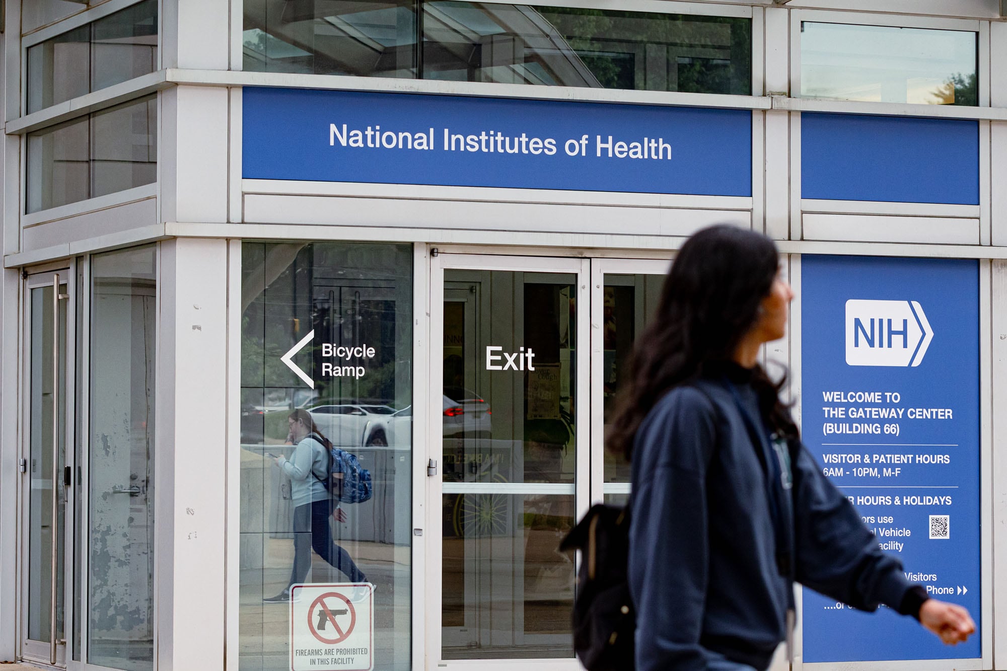 A photograph of a person walking in front of a building entry way. The sign above the entrance reads "National Institutes of Health."