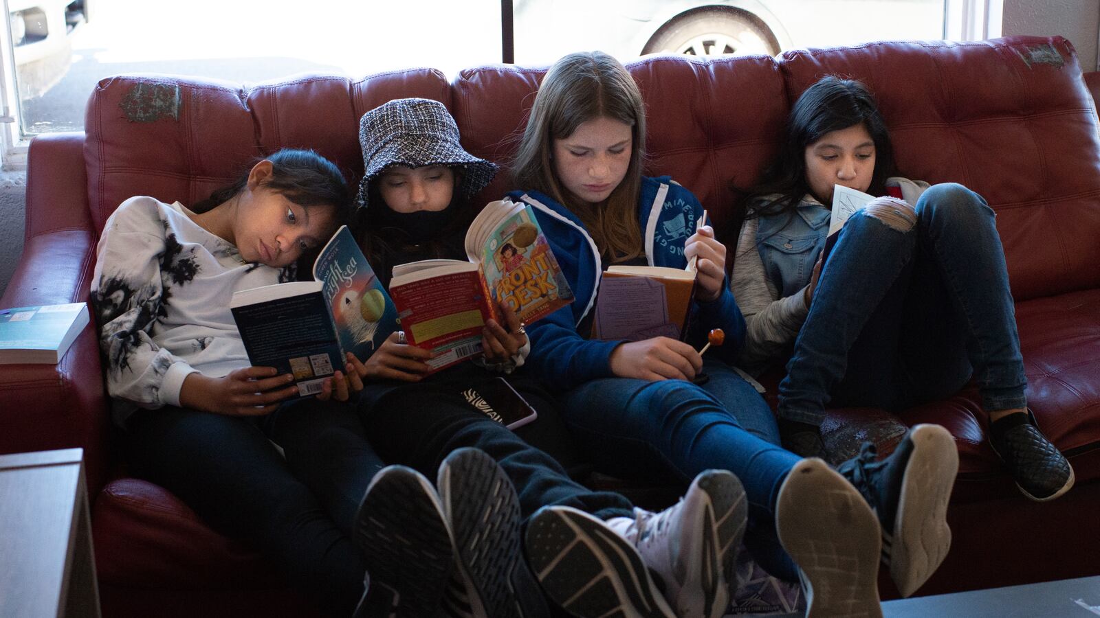 Four girls sit on a couch, their feet up on a coffee table, each reading their own book. They’re nestled together and look comfortable with each other.