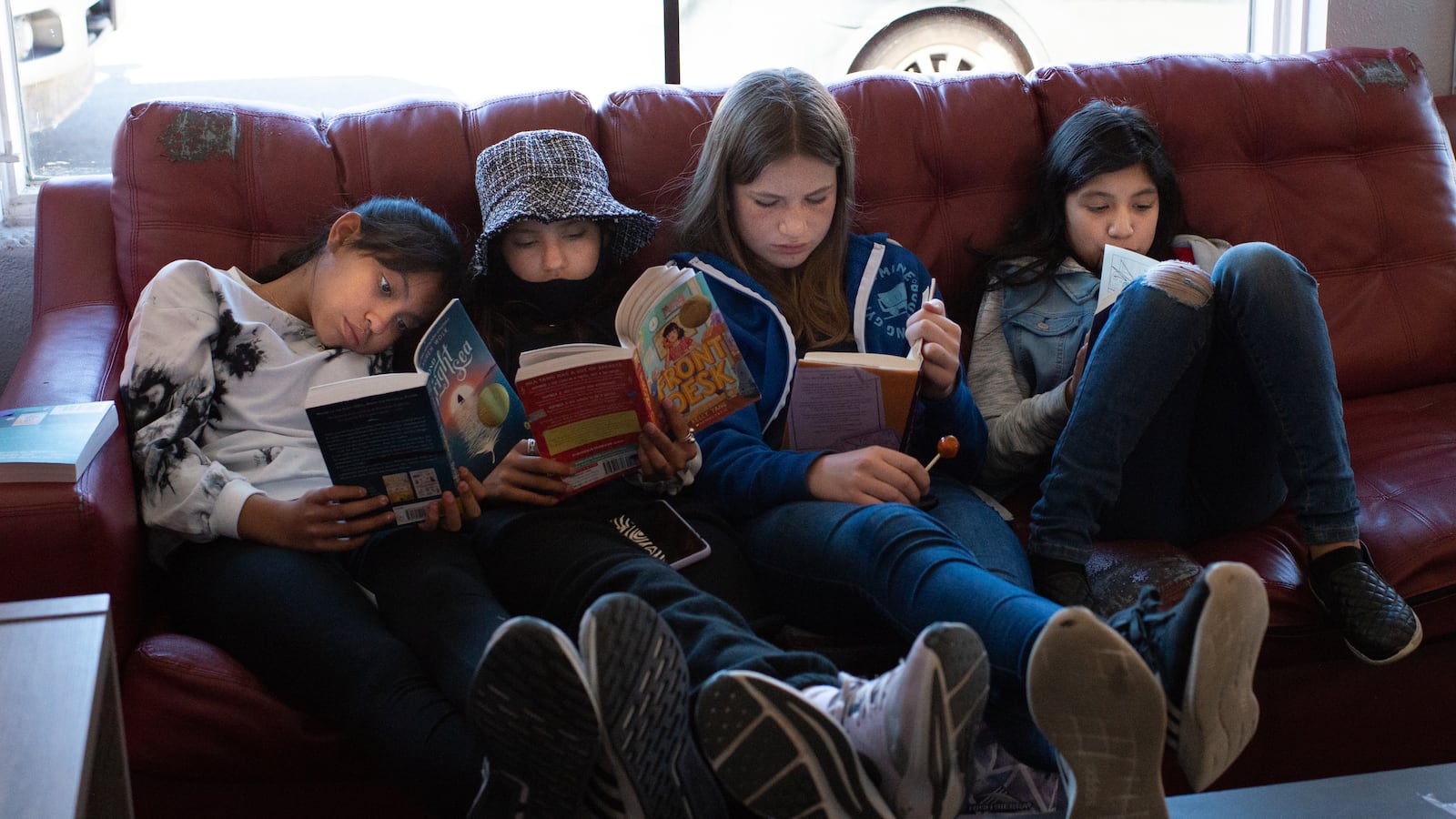 Four girls sit on a couch, their feet up on a coffee table, each reading their own book. They’re nestled together and look comfortable with each other. 