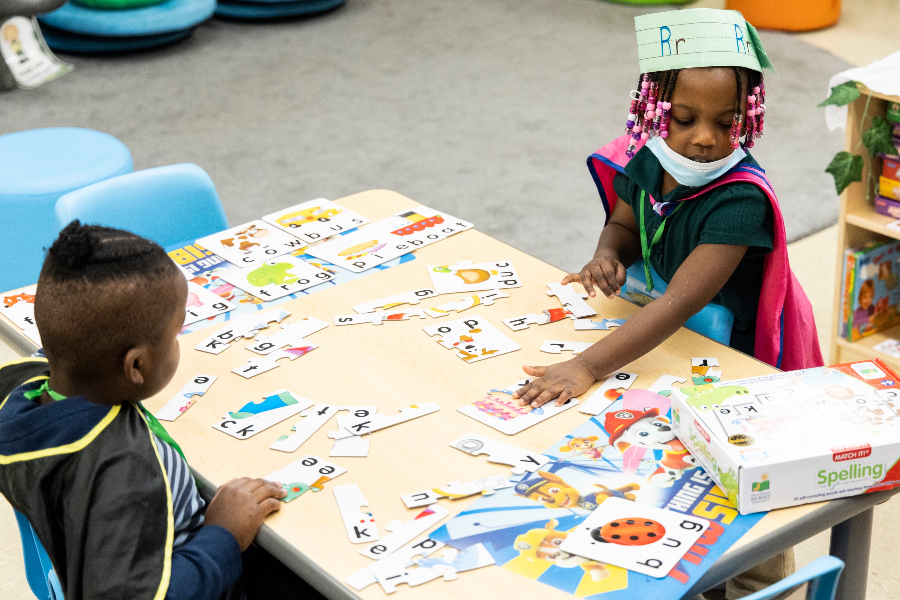 Two young children sit at a small wooden table with word puzzle pieces on top in a classroom.