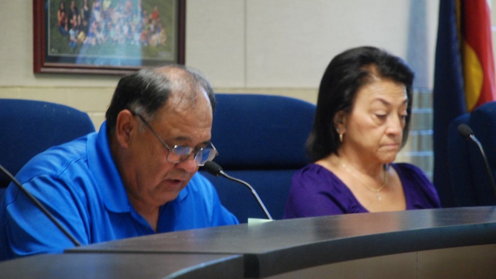 A man wearing glasses in a blue shirt and a woman also wearing a blue shirt sit at a table. Both are looking down.