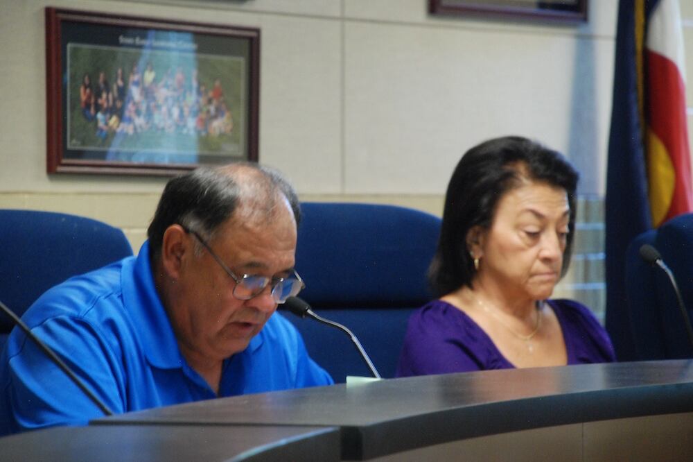 A man wearing glasses in a blue shirt and a woman also wearing a blue shirt sit at a table. Both are looking down.