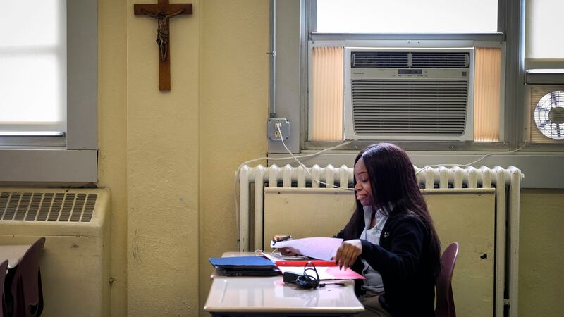 A cross hangs on a wall near a student looking through papers at a desk in a classroom in a Catholic school.