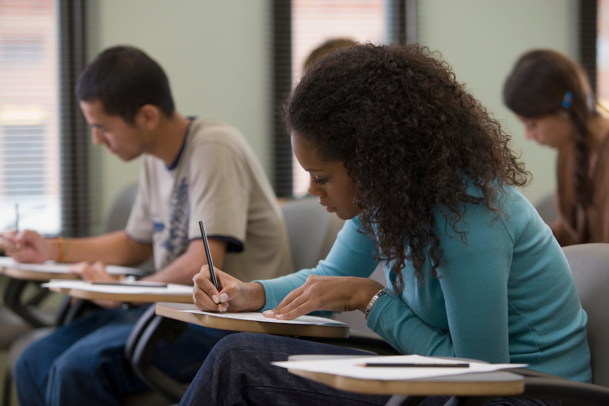 A student wearing a blue shirt with curly hair sits at a desk while holding a pencil and staring at a piece of paper.
