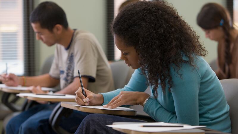 A student wearing a blue shirt with curly hair sits at a desk while holding a pencil and staring at a piece of paper.