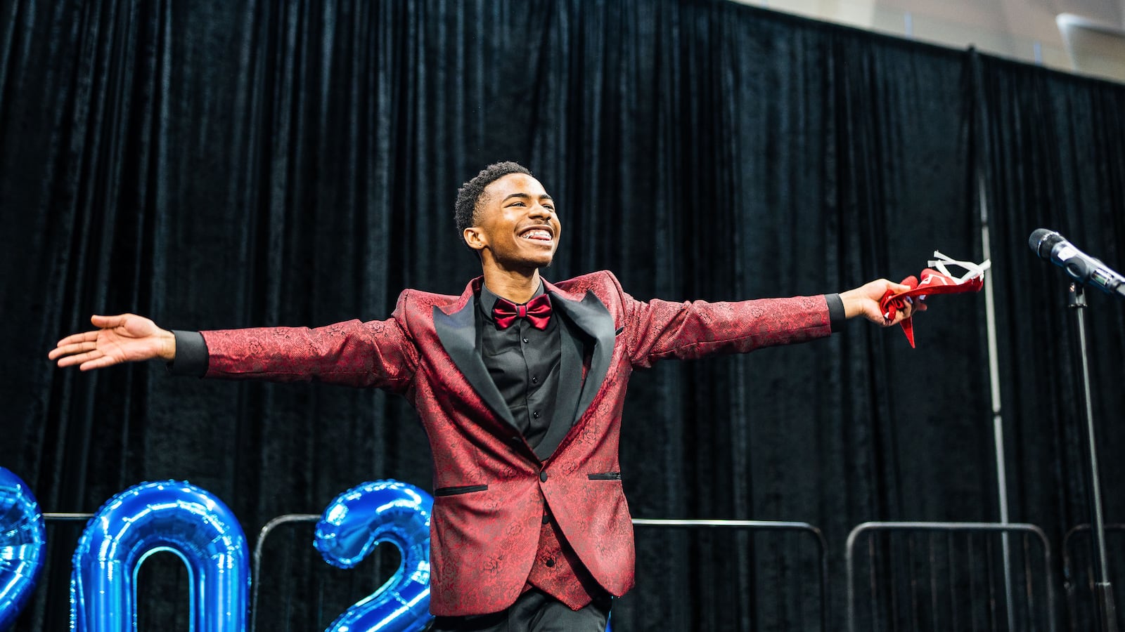 A young African American male smiles with his hands out to the side. He wears a maroon and black suit.