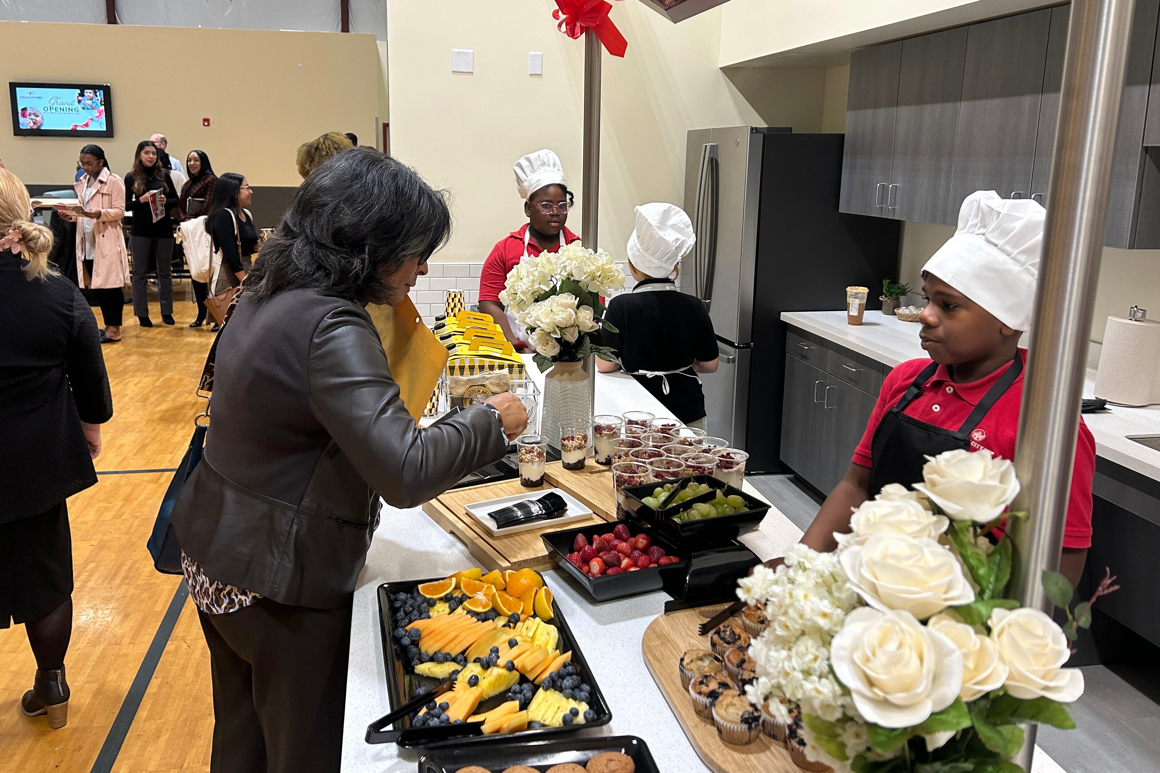 A woman in a dark jacket selects food from a counter. A student wearing a red shirt and a chef's hat watches her from a kitchen area on the other side of the counter.